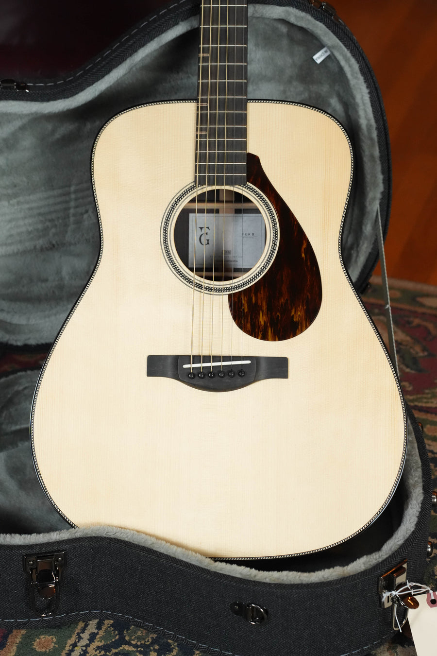A Yamaha FG9 R NT Adirondack/Indian Rosewood Dreadnought Acoustic Guitar by Yamaha rests in an open, gray-lined hard case on a patterned rug.