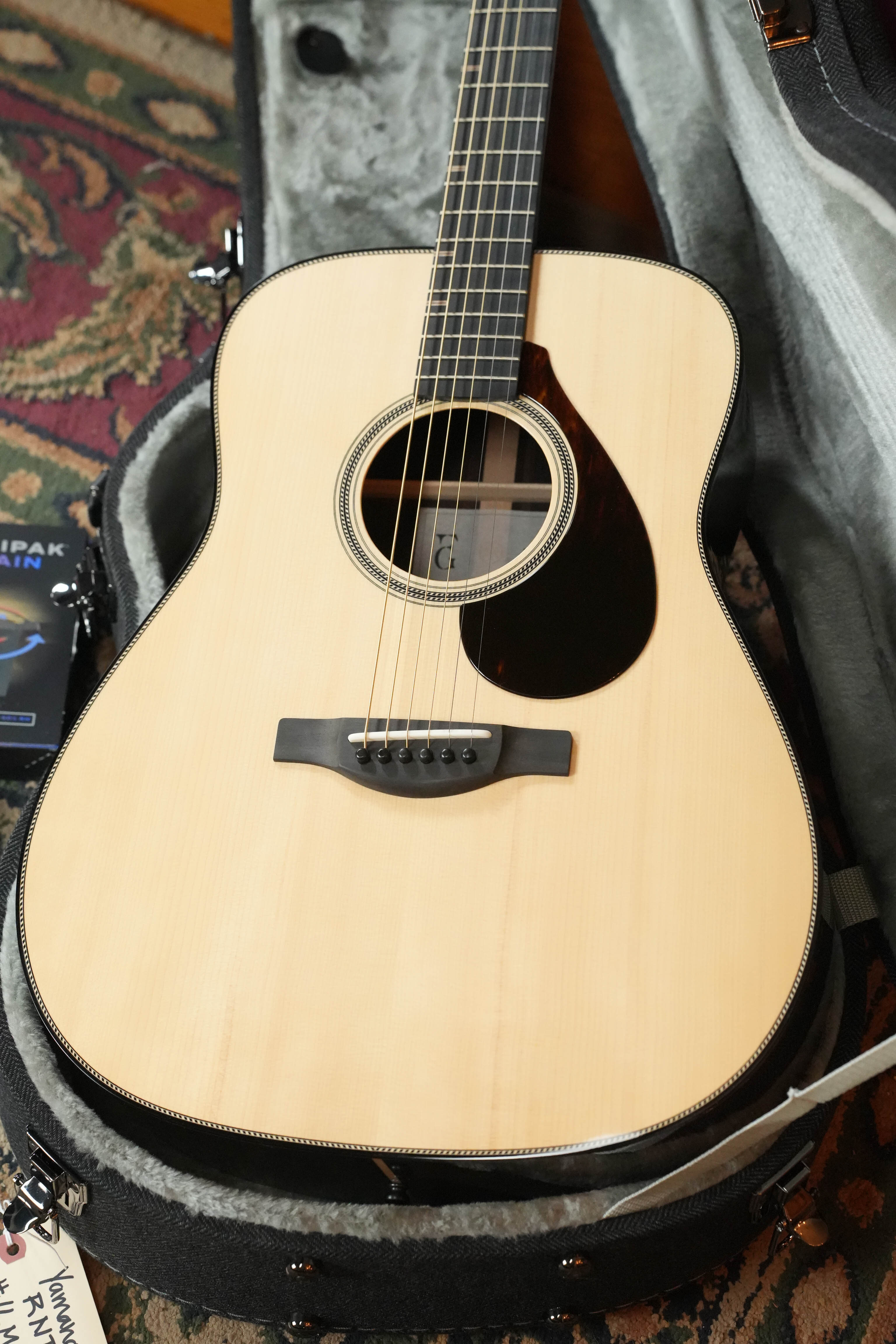 A Yamaha FG9 R NT Natural Finish Dreadnought Acoustic Guitar #126A rests in a plush, gray-lined hard case, with the open lid revealing its front and part of a colorful rug underneath.