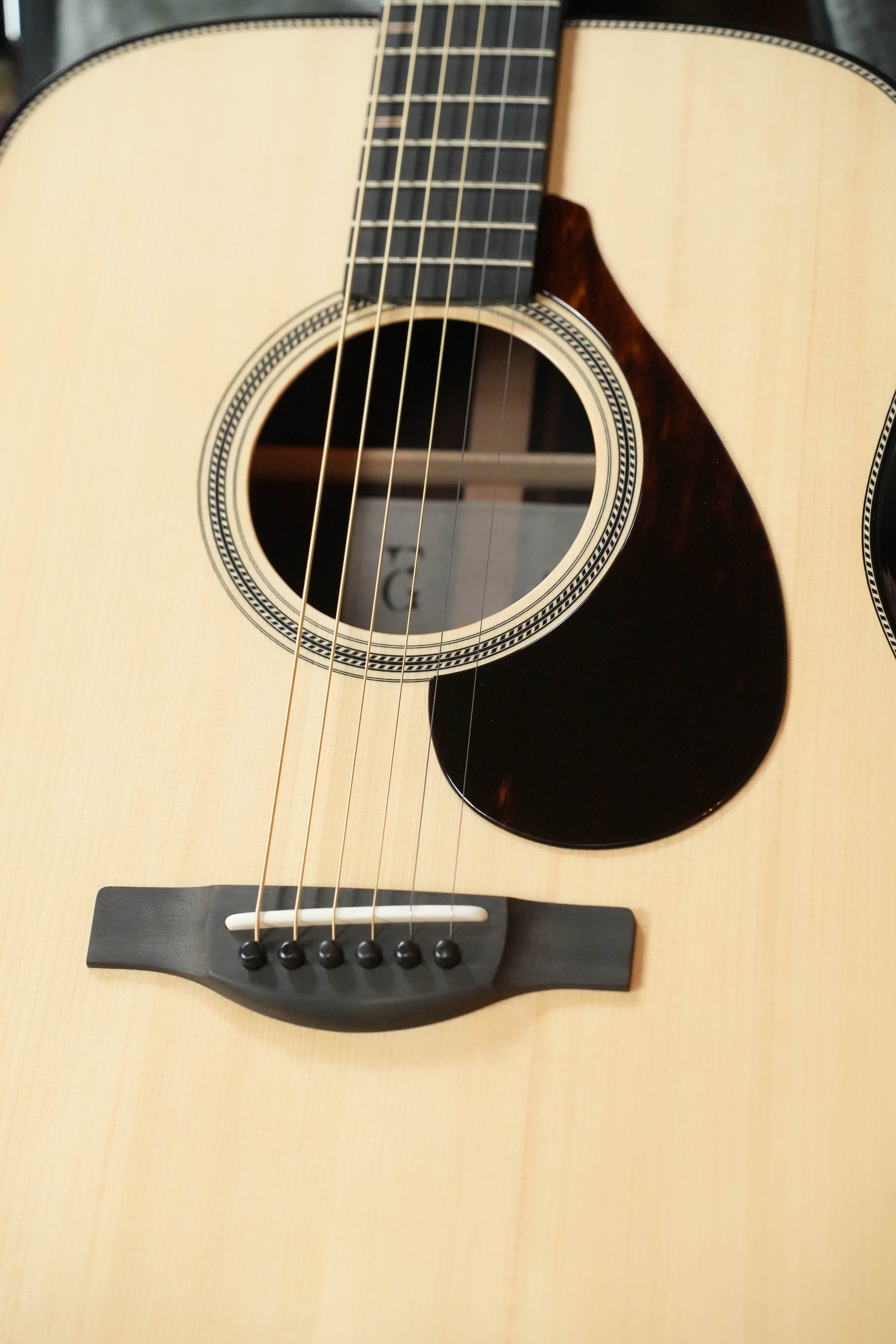 A close-up of the Yamaha FG9 R NT Natural Finish Dreadnought Acoustic Guitar #126A by Yamaha shows its Adirondack spruce body, sound hole, black pickguard, six-pin bridge, and strings against a neutral background.