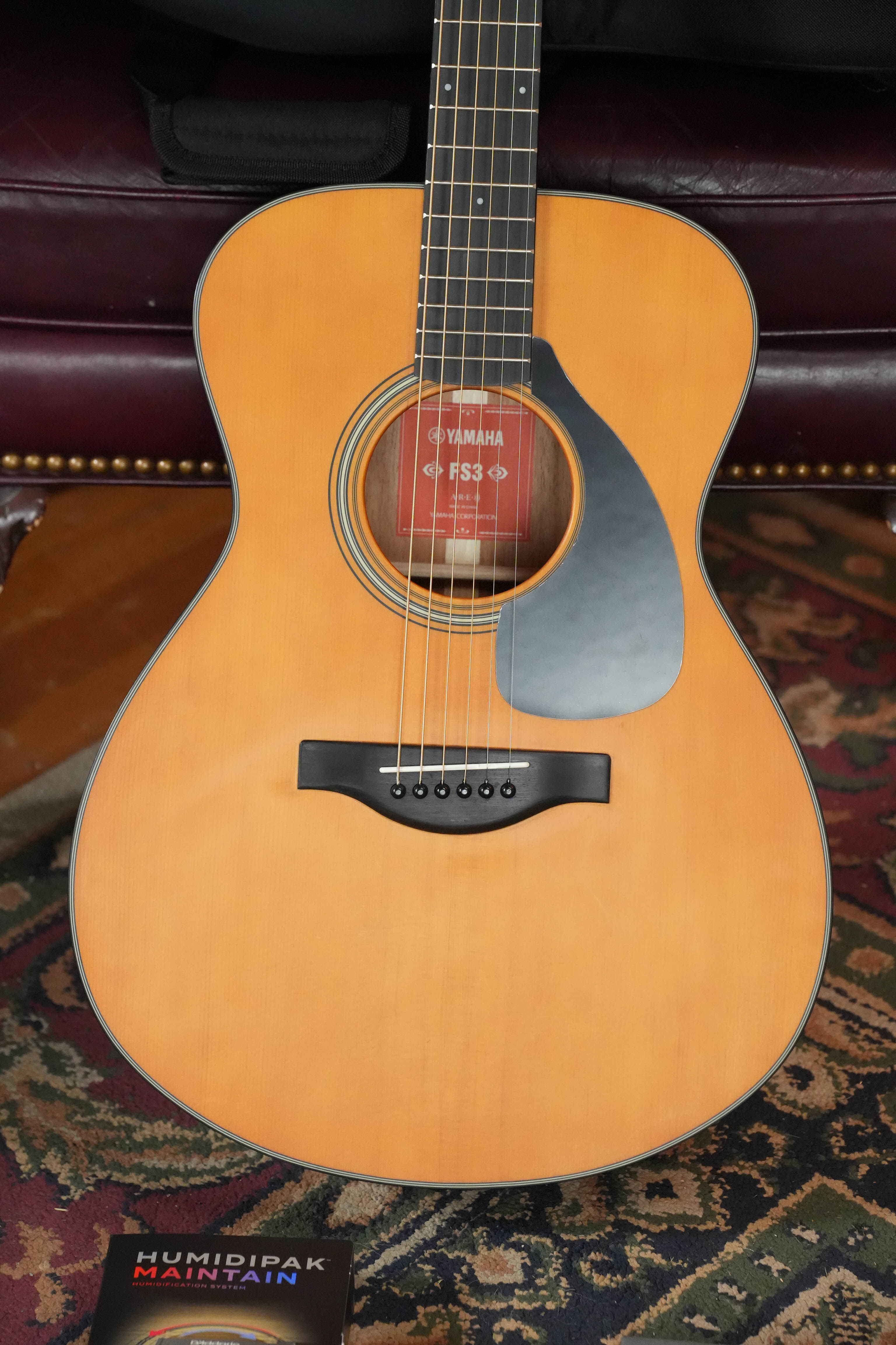 A Yamaha FS3 Red Label Concert Acoustic Guitar #1516 with a natural wood finish rests on a patterned rug, accompanied by a Humidipak maintenance kit.