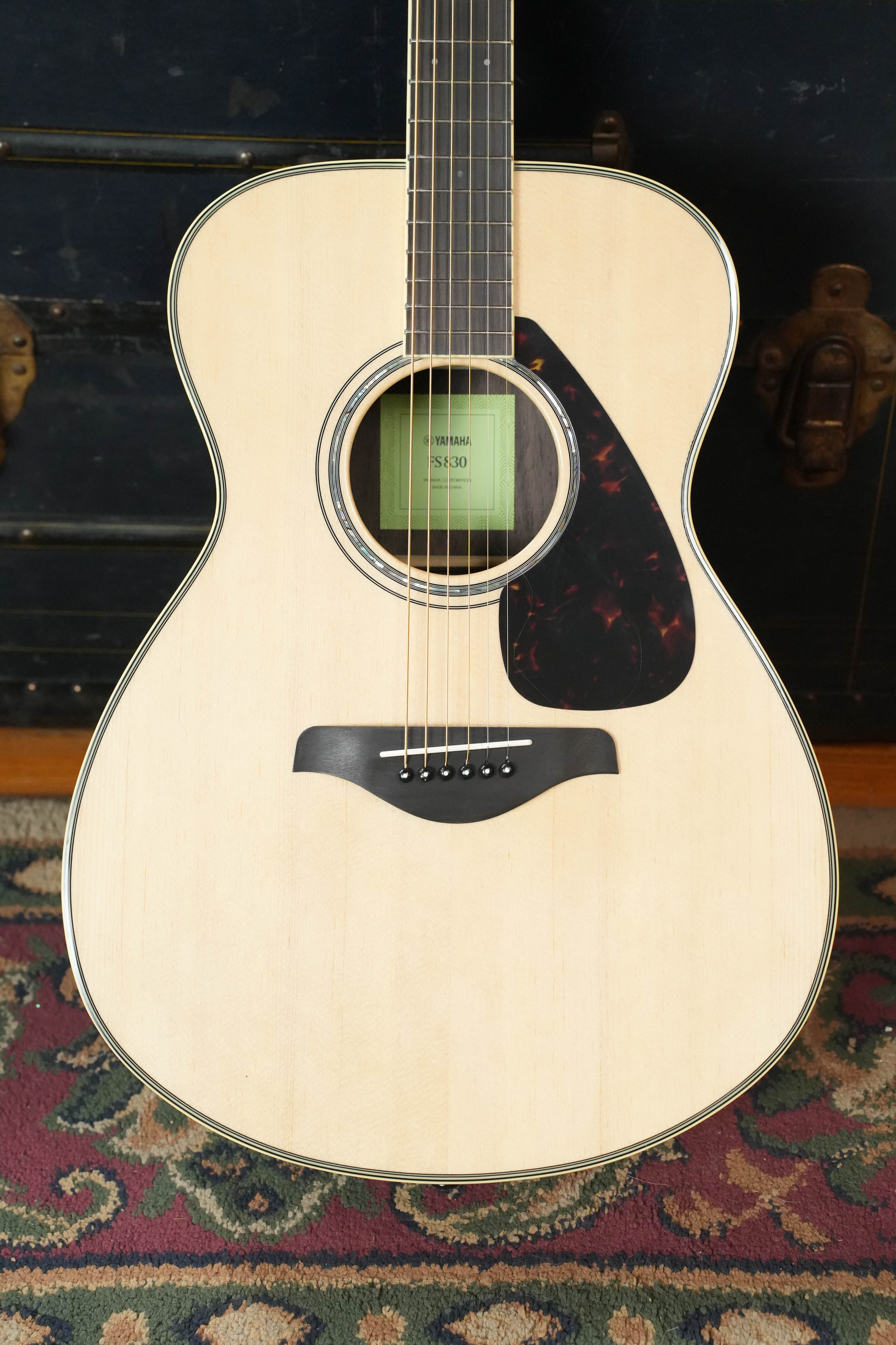 A Yamaha FS830 Concert Acoustic Guitar #1751 by Yamaha, featuring a solid spruce top, light body, and black pickguard, stands upright on a patterned rug against a dark wooden background.