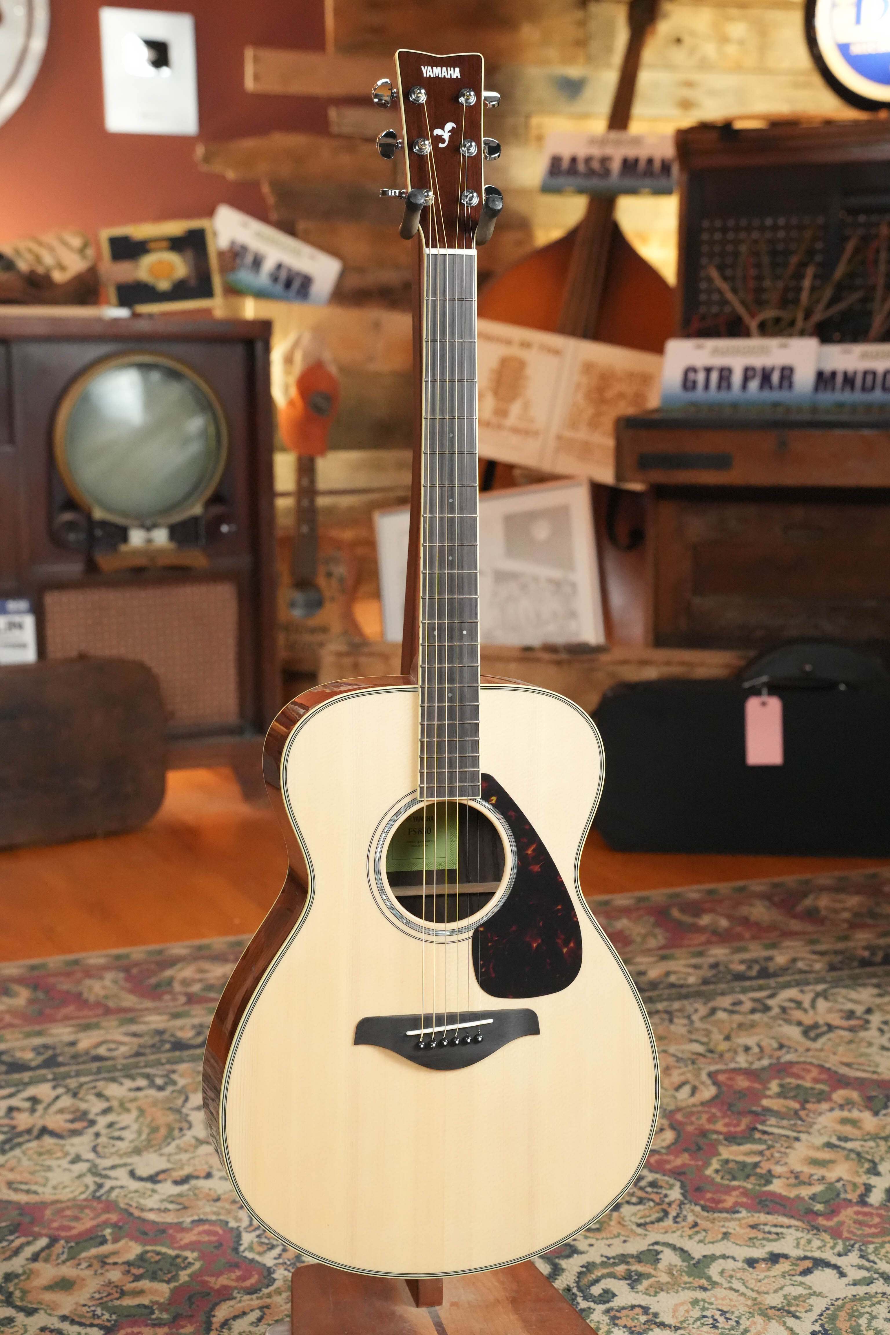 The Yamaha FS830 Concert Acoustic Guitar #1751 by Yamaha, featuring a solid spruce top and natural finish, stands upright on a patterned rug in a cozy vintage-style room filled with musical instruments and retro decor.