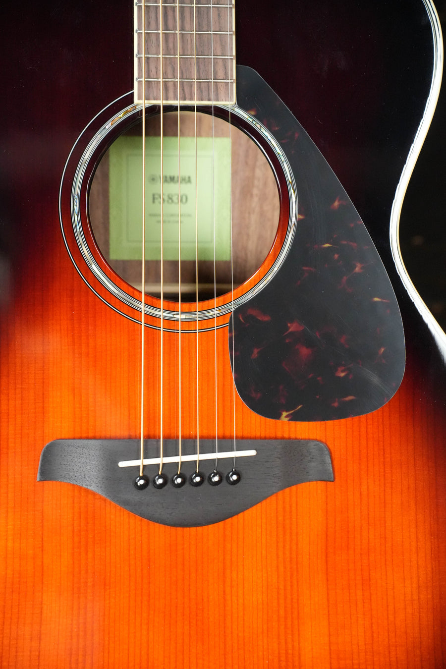 Close-up of a Yamaha FS830 TBS Tobacco Sunburst Concert Acoustic Guitar #0129 body, showing six strings, black pickguard, wooden bridge, solid spruce top, and the guitar label inside the soundhole. Brand: Yamaha.