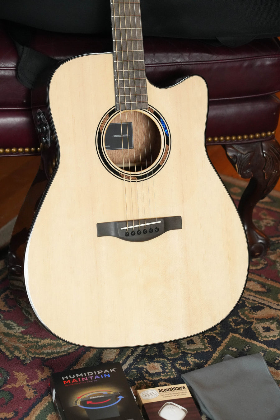 A Yamaha TAG1 C Natural Finish Dreadnought Cutaway Acoustic-Electric Guitar rests on a patterned rug before a dark leather chair, with accessories like a Humidpak and polishing cloth placed nearby.