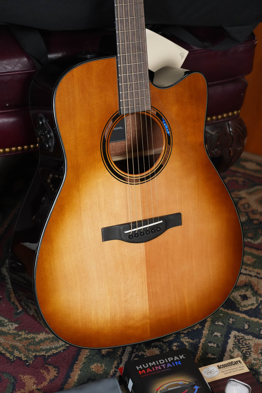 A Yamaha TAG1 C Sitka/Mahogany Sandburst Dreadnought Transacoustic Guitar w/Cutaway (#0610) from Yamaha rests on a dark leather surface beside a Humidipak Maintenance packet, placed atop a patterned rug.