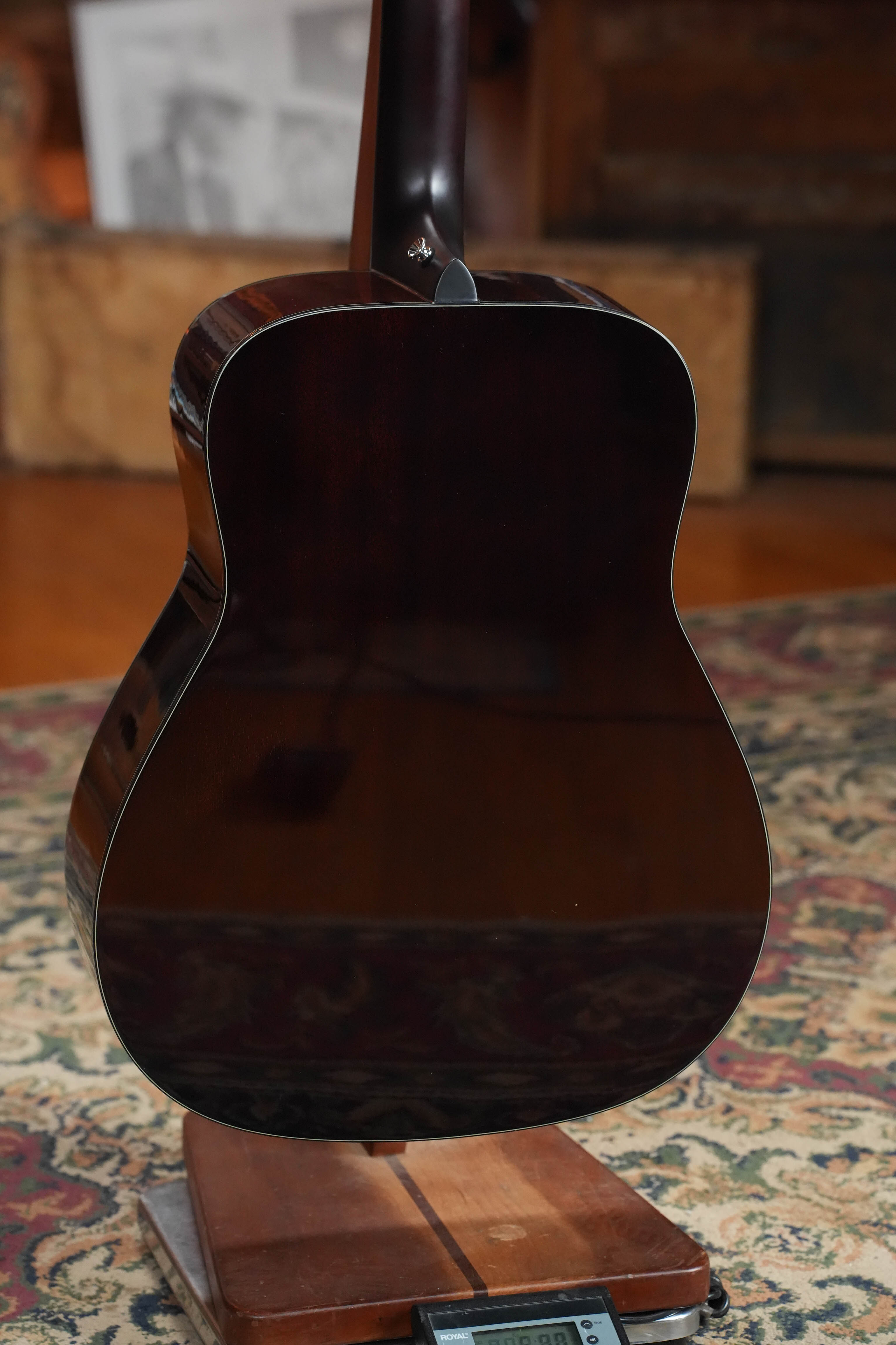 A Yamaha TAG1E TBL Spruce/Mahogany Transacoustic Dreadnought Guitar #1280 is shown from the back on a wooden stand atop a patterned rug, with blurred boxes and papers in the background.