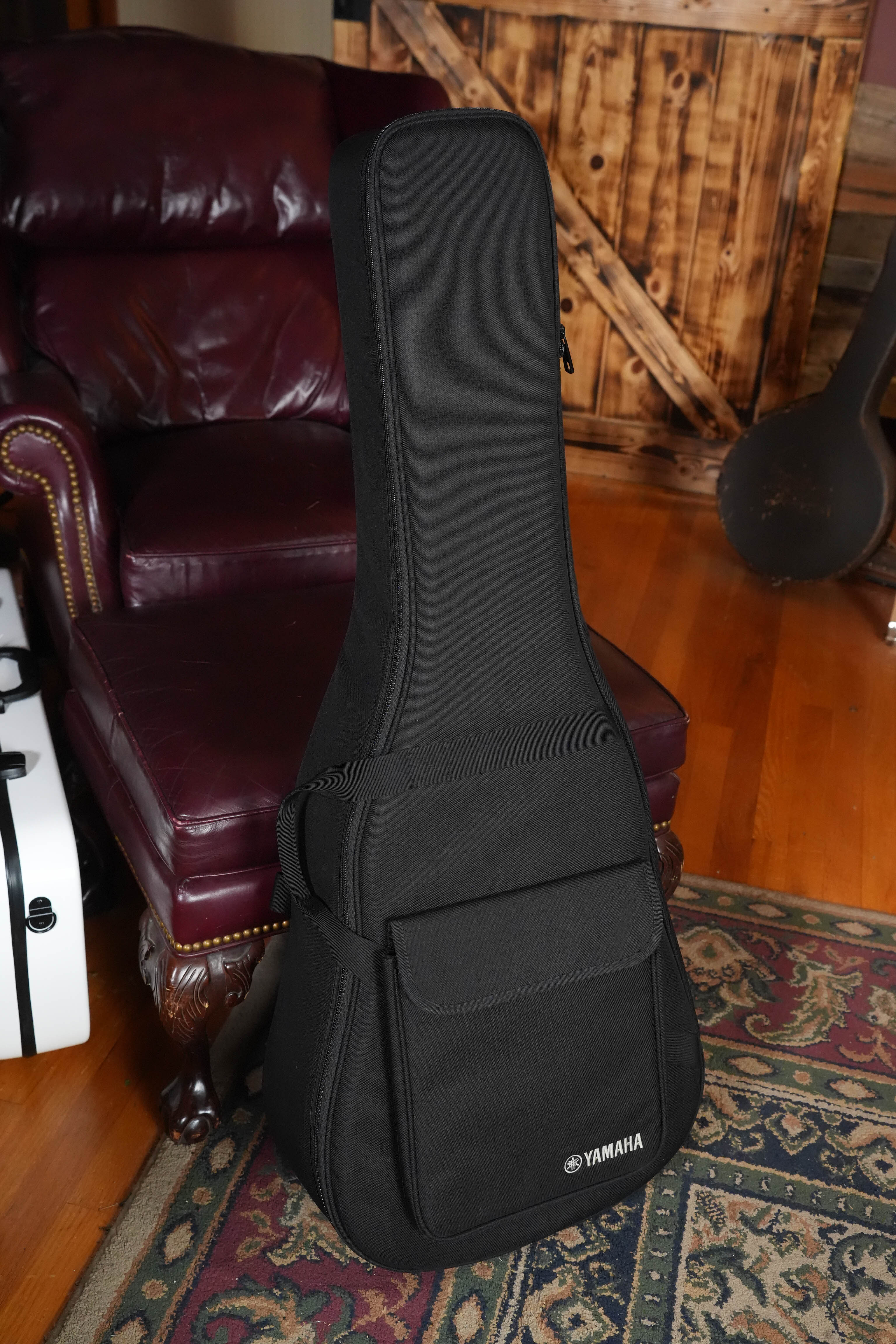 A Yamaha TAG1E TBL Transacoustic Dreadnought Guitar stands upright in its gig bag on a patterned rug beside a maroon leather armchair, set in a cozy room with wooden floors and rustic wooden doors.