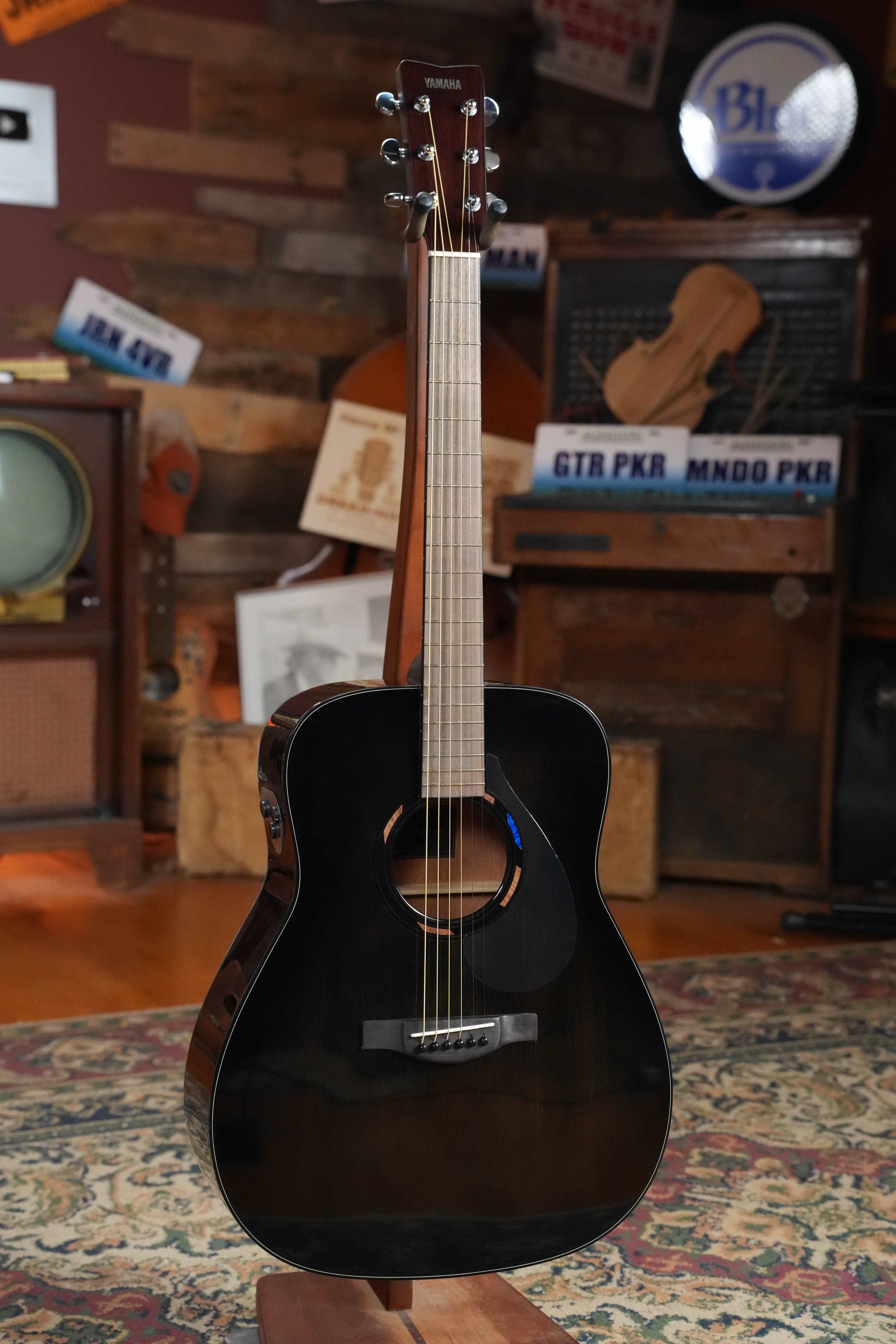 A Yamaha TAG1E TBL Spruce/Mahogany TransAcoustic Dreadnought Guitar #1280 stands upright on a patterned rug, surrounded by vintage radios, music-themed decor, and wooden crates in the background.