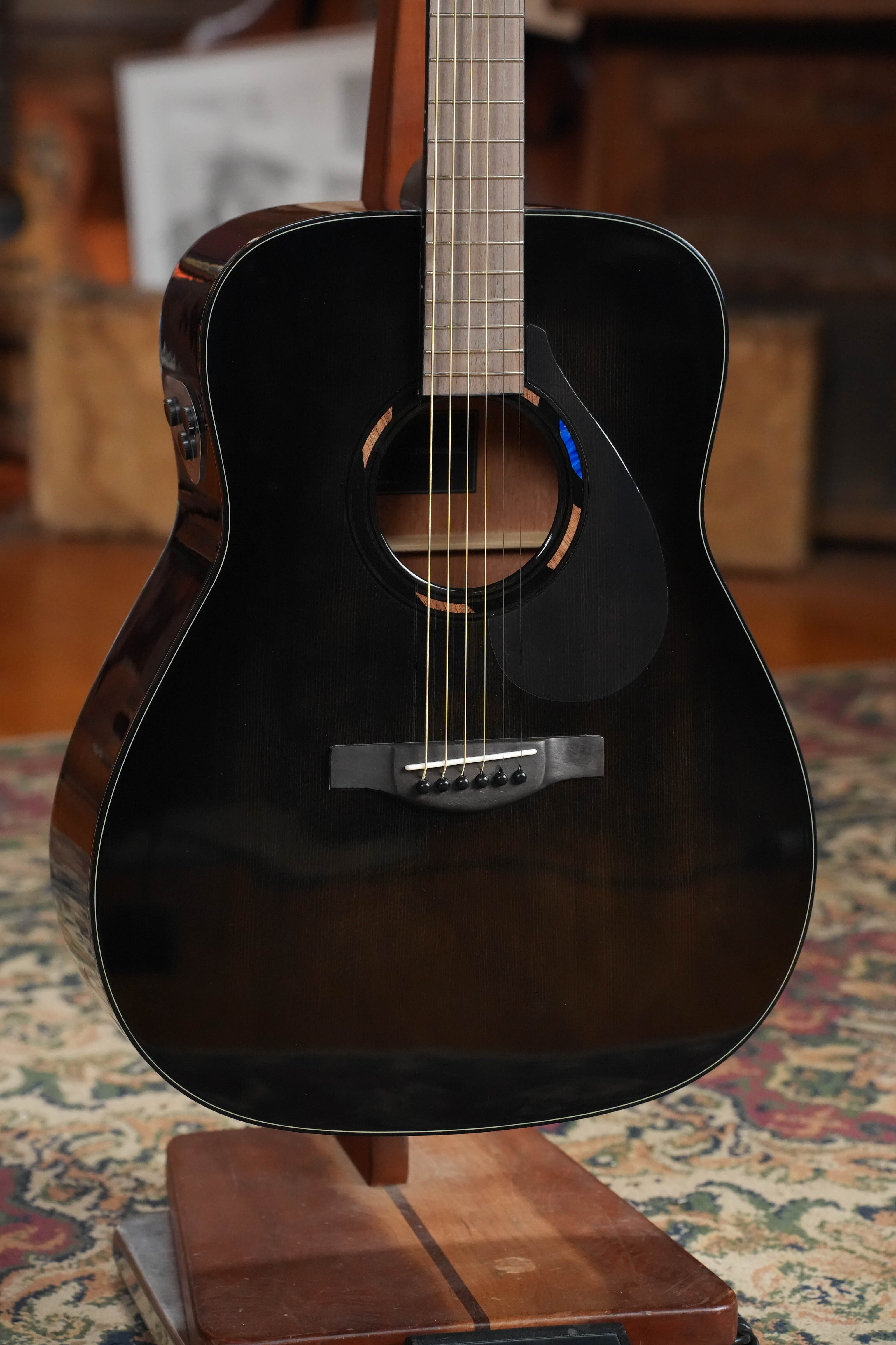 A Yamaha TAG1E TBL Spruce/Mahogany Transacoustic Dreadnought Guitar (#1280) with built-in reverb and chorus rests on a wooden stand, its fretboard and sound hole partially visible against a patterned rug and blurred indoor setting.