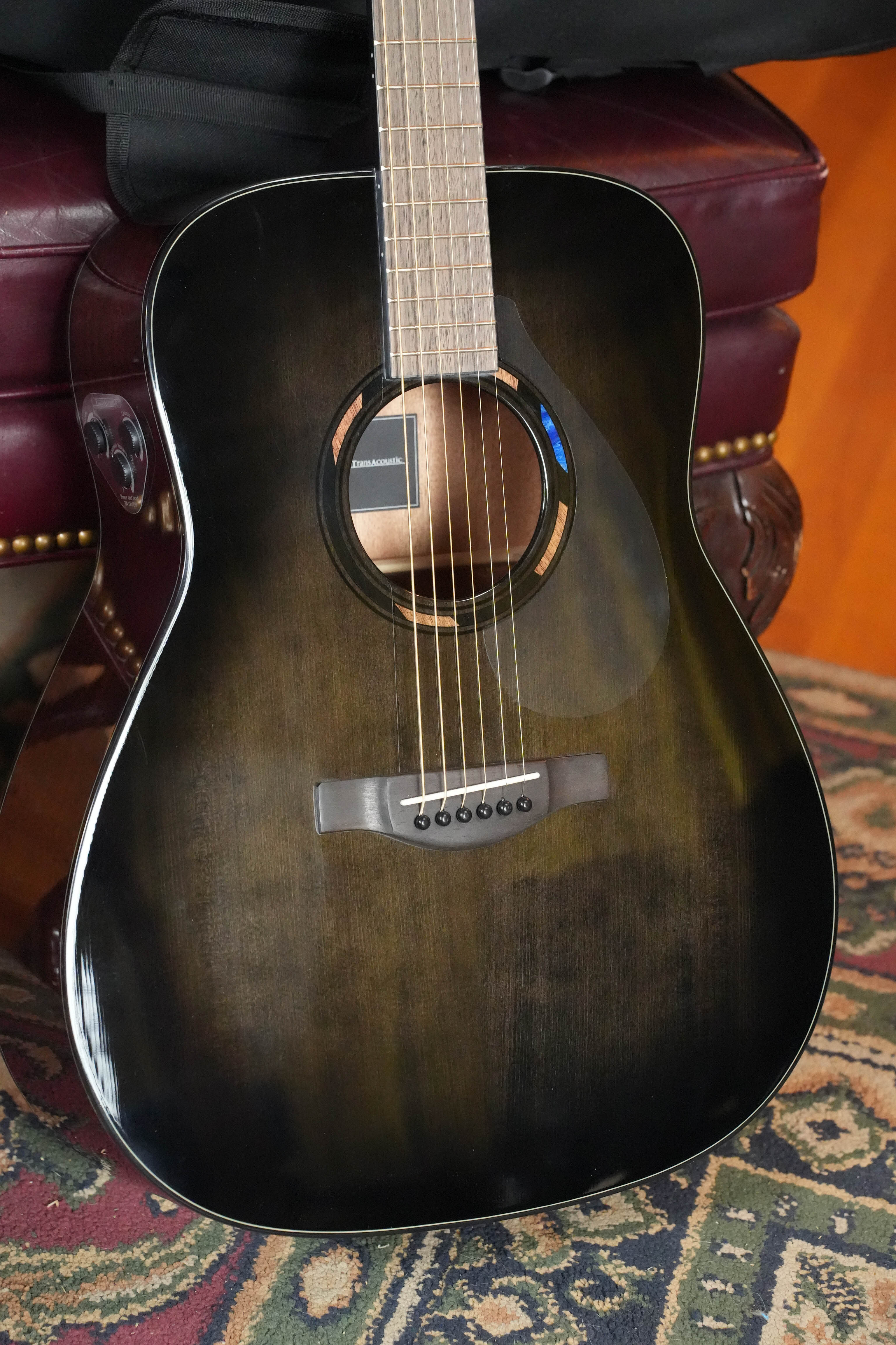 A Yamaha TAG1E TBL TransAcoustic Translucent Black Finish Dreadnought Acoustic-Electric Guitar #1164 sits on a burgundy leather chair with gold studs, atop a patterned rug.