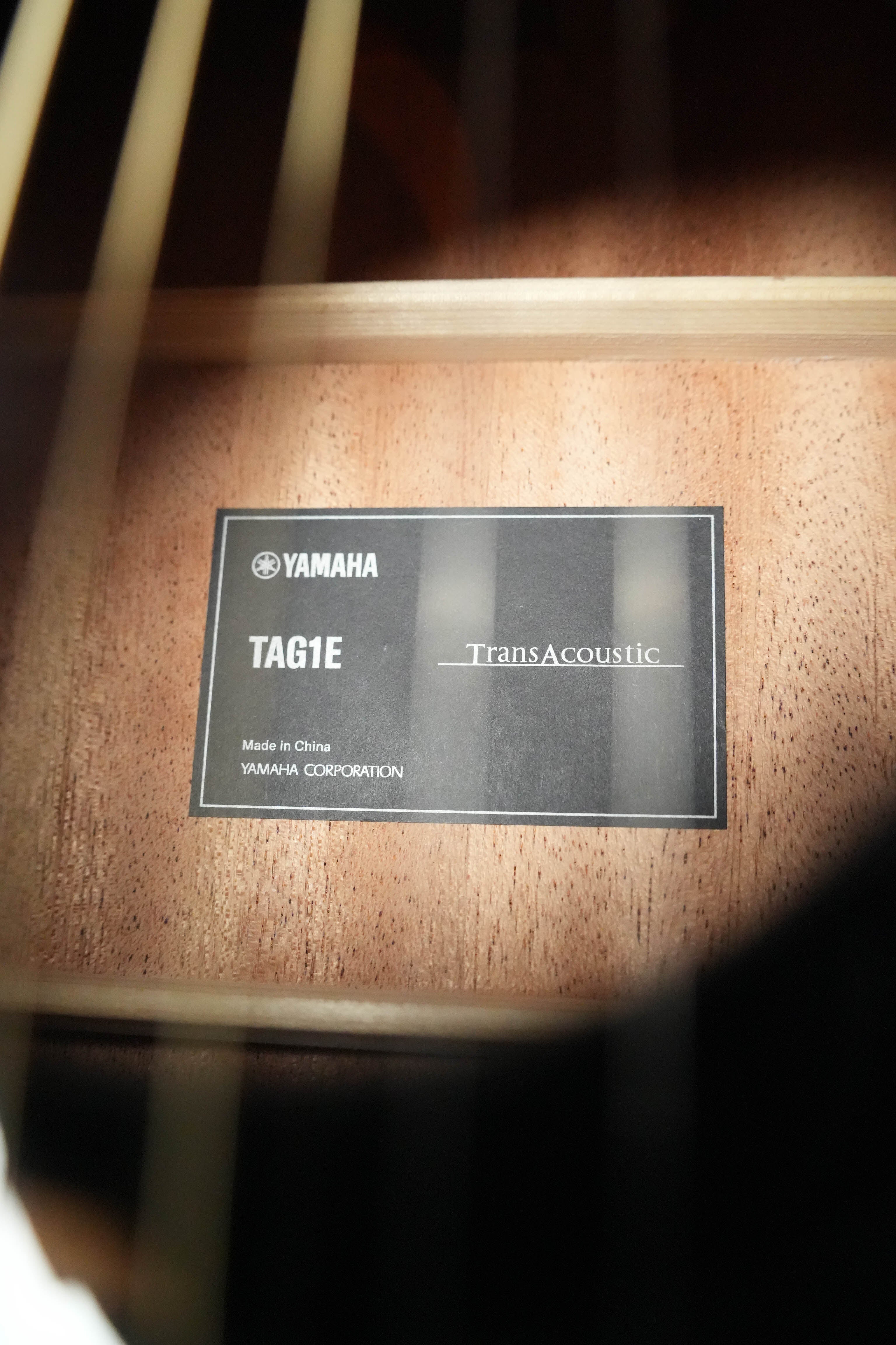 Close-up inside a Yamaha TAG1E TBL TransAcoustic Dreadnought Acoustic-Electric Guitar #1164, showing the label with TransAcoustic system details, Made in China and Yamaha Corporation. Wood grain is visible in the background.
