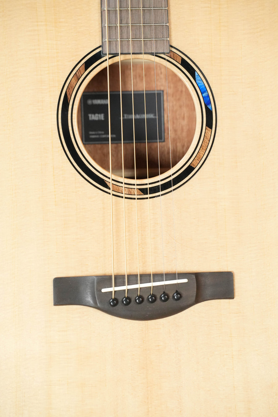 Close-up of the Yamaha TAG1E TransAcoustic Natural Finish Dreadnought Acoustic-Electric Guitar #1116, highlighting its sound hole, strings, internal label, and dark wood bridge with six pins—showcasing Yamaha's classic craftsmanship.