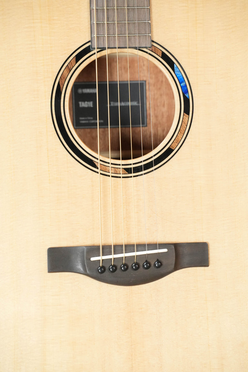 Close-up of the Yamaha TAS1E Natural Finish Concert Acoustic-Electric Guitar #1133 by Yamaha, showing its solid spruce top, mahogany back and sides, sound hole, strings, bridge, fretboard, and visible Yamaha label.
