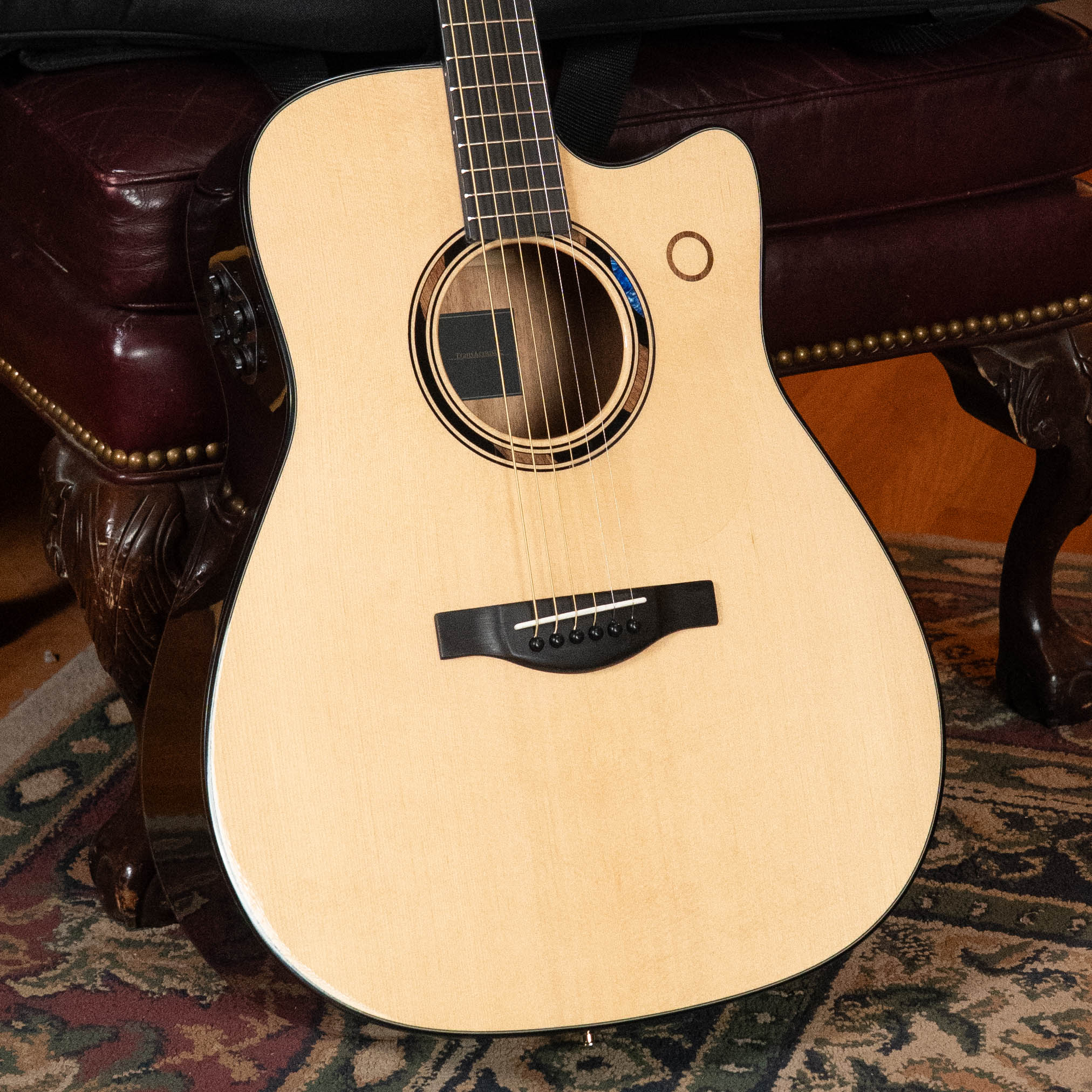 A Yamaha TAG3C-NT Sitka/Mahogany Natural Dreadnought Guitar with the newest TransAcoustic System (#0558) rests on a patterned rug by a dark leather chair, highlighting built-in effects for enhanced sound.