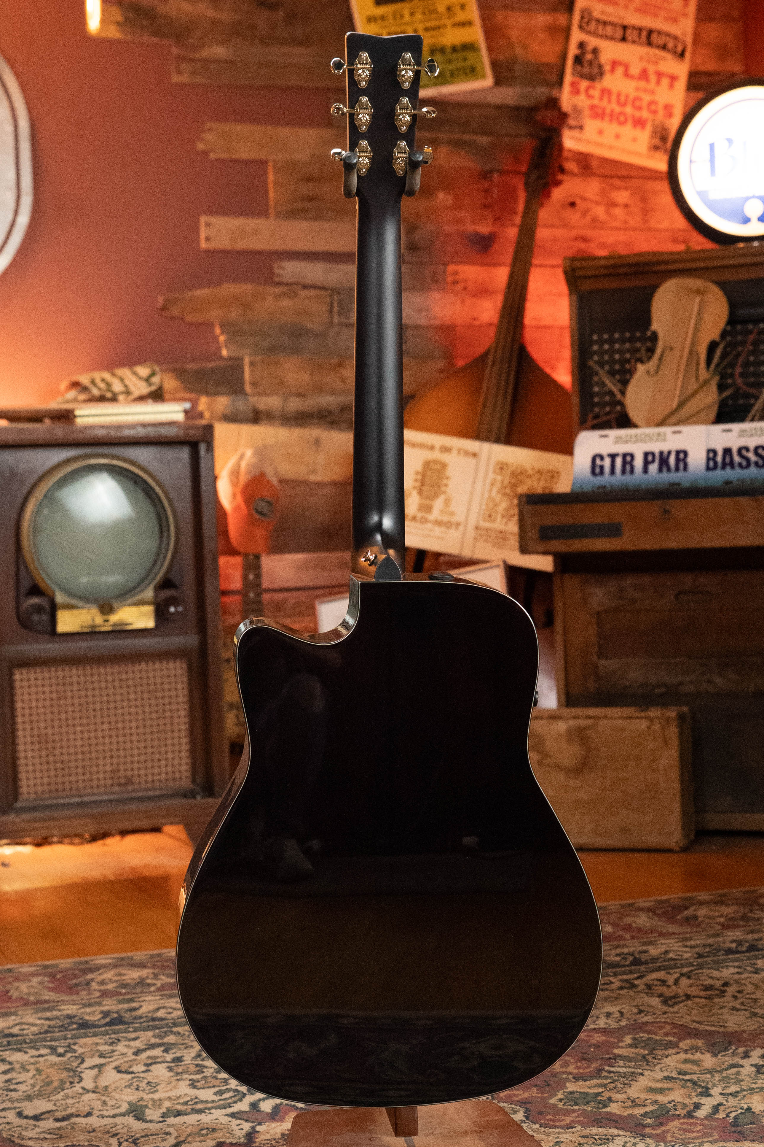 A Yamaha TAG3C-NT Natural Dreadnought Guitar with the latest TransAcoustic System is displayed upright from the back in a cozy vintage room with wooden walls, retro signs, an old TV, and musical instruments in the background.