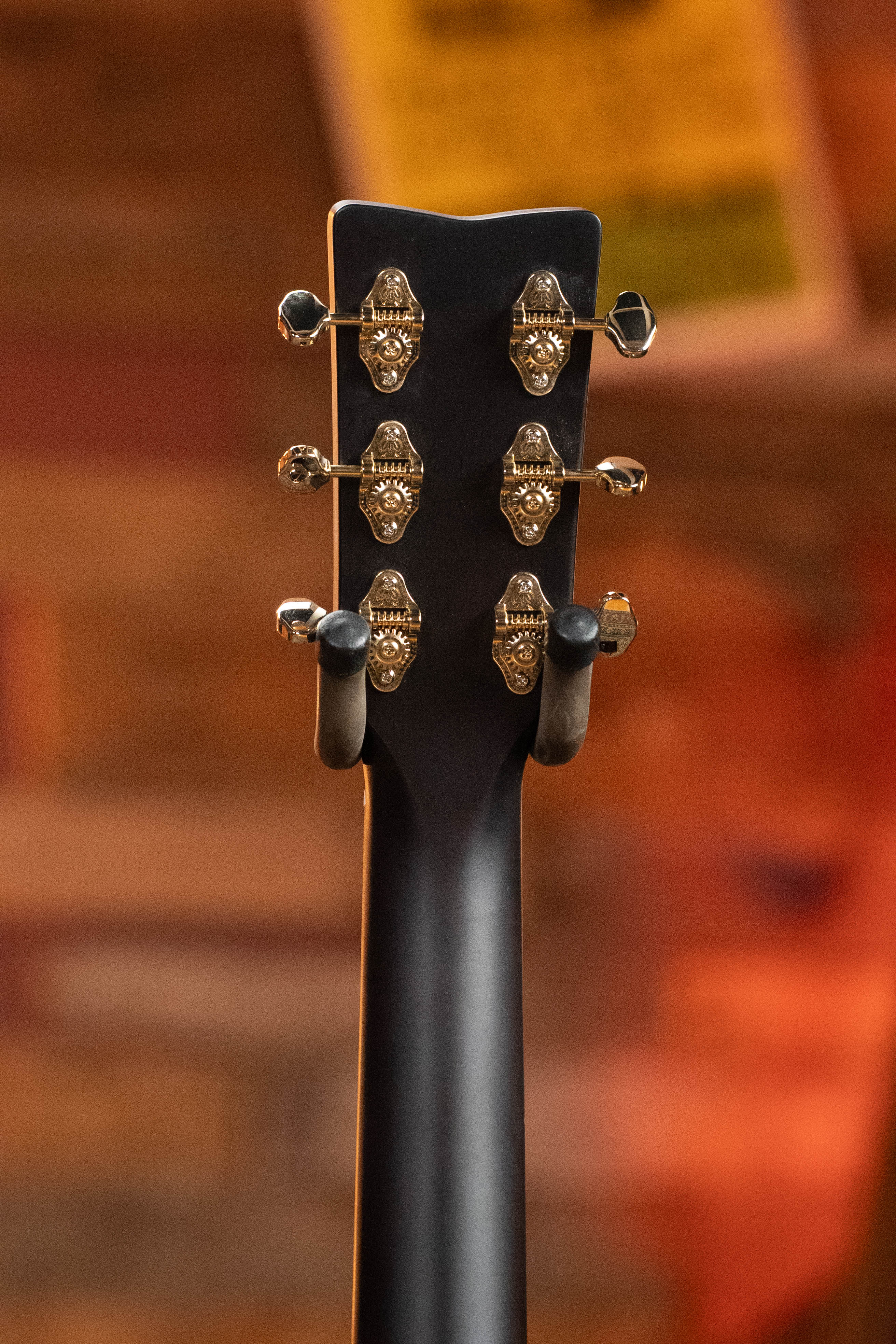 The headstock of a Yamaha TAG3C-NT Sitka/Mahogany Natural Dreadnought Guitar with six gold tuners is shown on a wall hanger, against a blurred warm brown and orange background.
