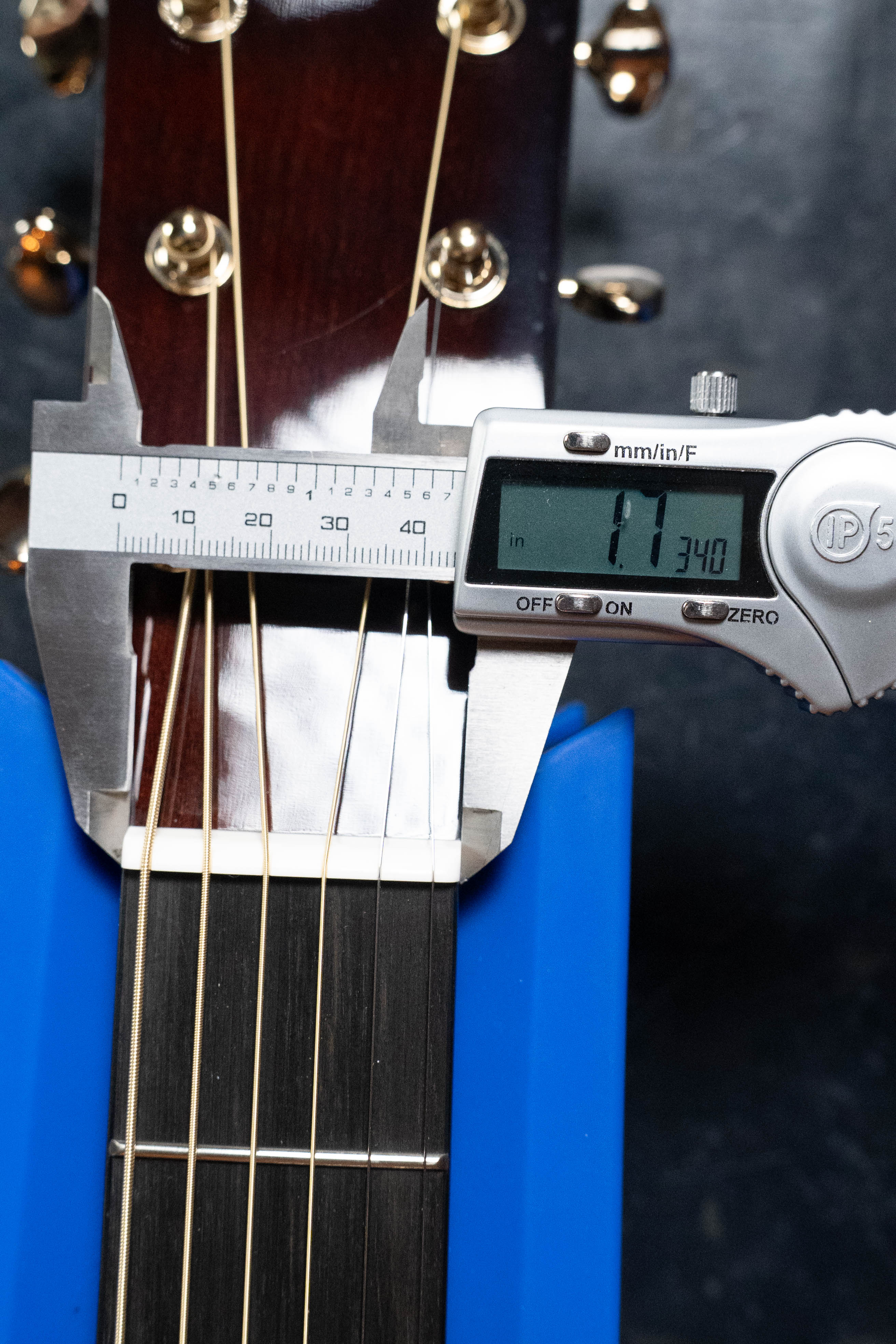 A digital caliper measures the nut width of a Yamaha TAG3C-NT Sitka/Mahogany Natural Dreadnought Guitar w/ TransAcoustic System, displaying 1.1340 inches. The guitar rests securely in a blue stand.