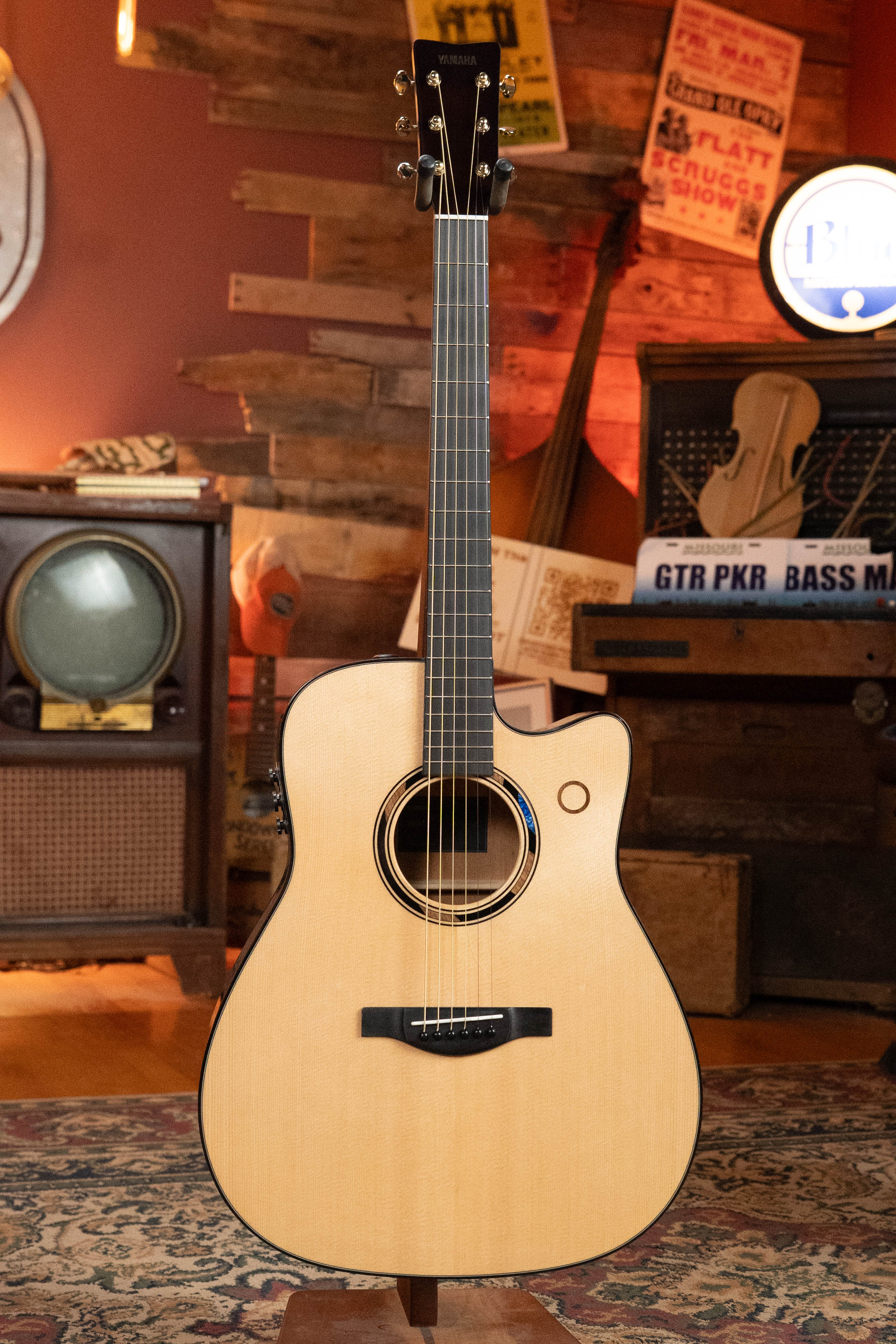 A Yamaha TAG3C-NT Sitka/Mahogany Natural Dreadnought Guitar with the newest TransAcoustic System is displayed on a stand in a cozy, vintage-style room with musical instruments, posters, and a TV in the background.