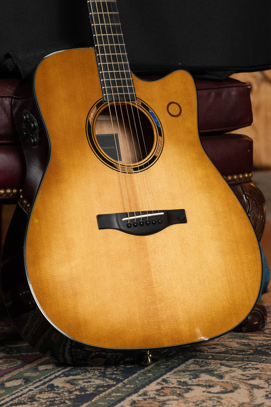 A close-up of a Yamaha TAG3C-SDB Sitka/Mahogany Sandburst Dreadnought Guitar with the NEWEST TransAcoustic System (#0426), resting upright against a dark brown leather chair on a patterned rug.