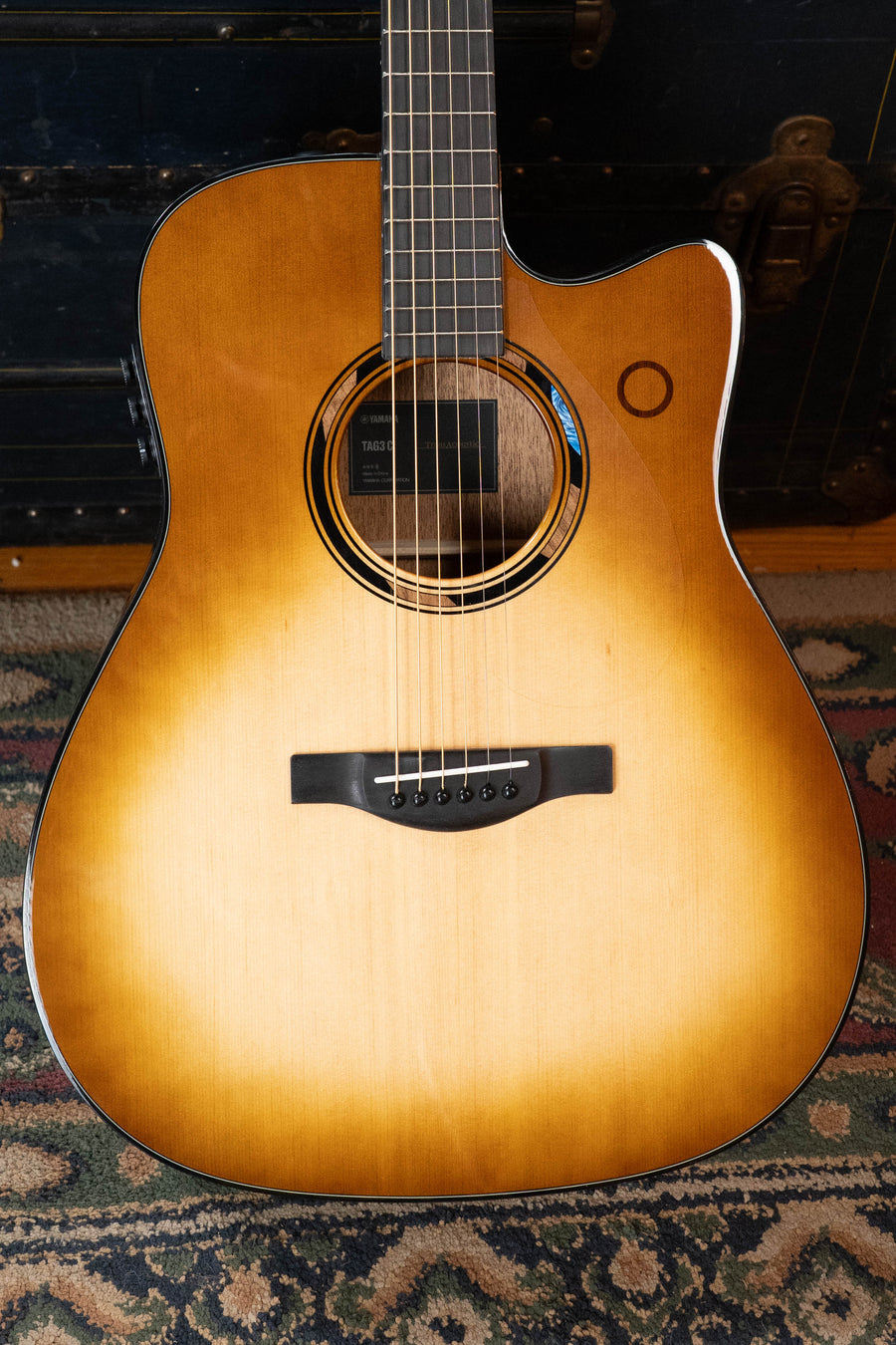 A Yamaha TAG3C-SDB Sitka/Mahogany Sandburst Dreadnought Guitar with the newest TransAcoustic System (#0592) rests on a patterned rug, cutaway body visible and a dark trunk in the background.