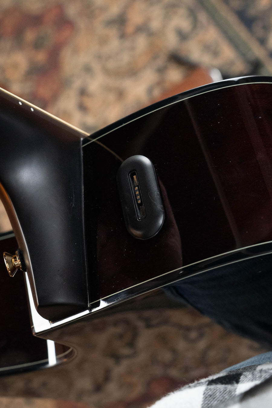 Close-up of a Yamaha TAG3C-SDB Sitka/Mahogany Sandburst Dreadnought Guitar, showing its electronic connector port and glossy finish. The NEWEST TransAcoustic System #0592 is featured, with a patterned rug and plaid shirt in the background.