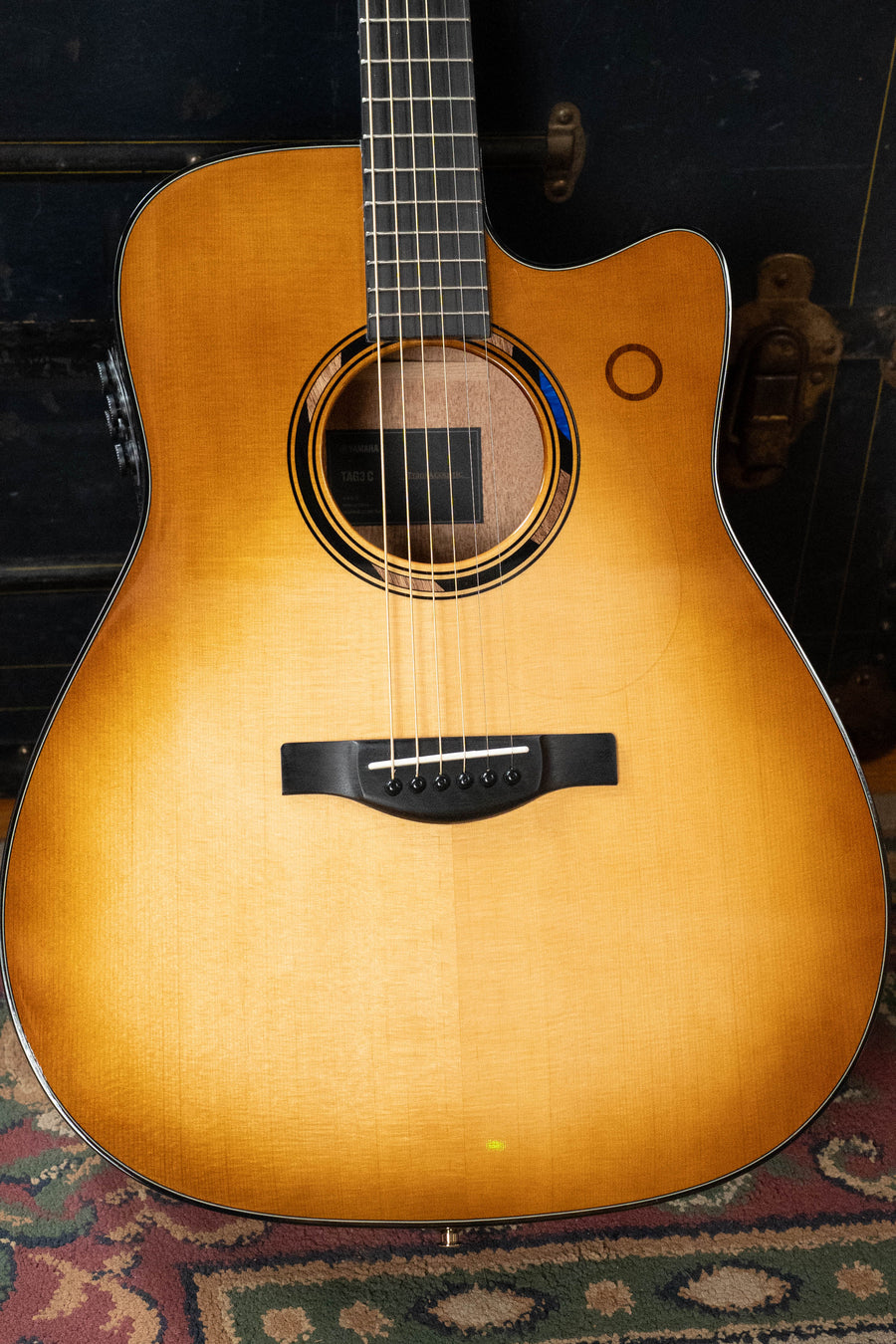 A Yamaha TAG3C-SDB Sandburst Dreadnought Guitar with Sitka/Mahogany body, decorative rosette, six strings, cutaway design, and the NEWEST TransAcoustic System rests on a patterned rug.