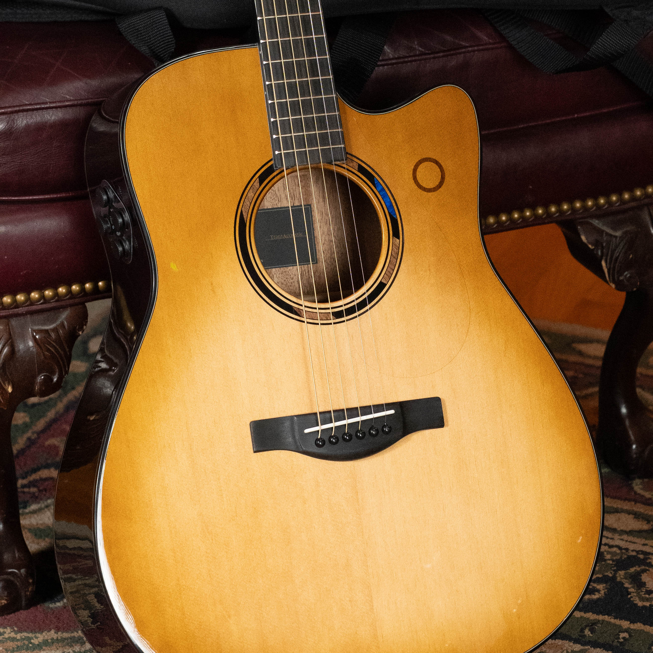 A Yamaha TAG3C-SDB Sitka/Mahogany Sandburst Dreadnought Guitar with the newest TransAcoustic System rests against a dark red leather chair above a patterned rug, featuring a cutaway design and six strings.