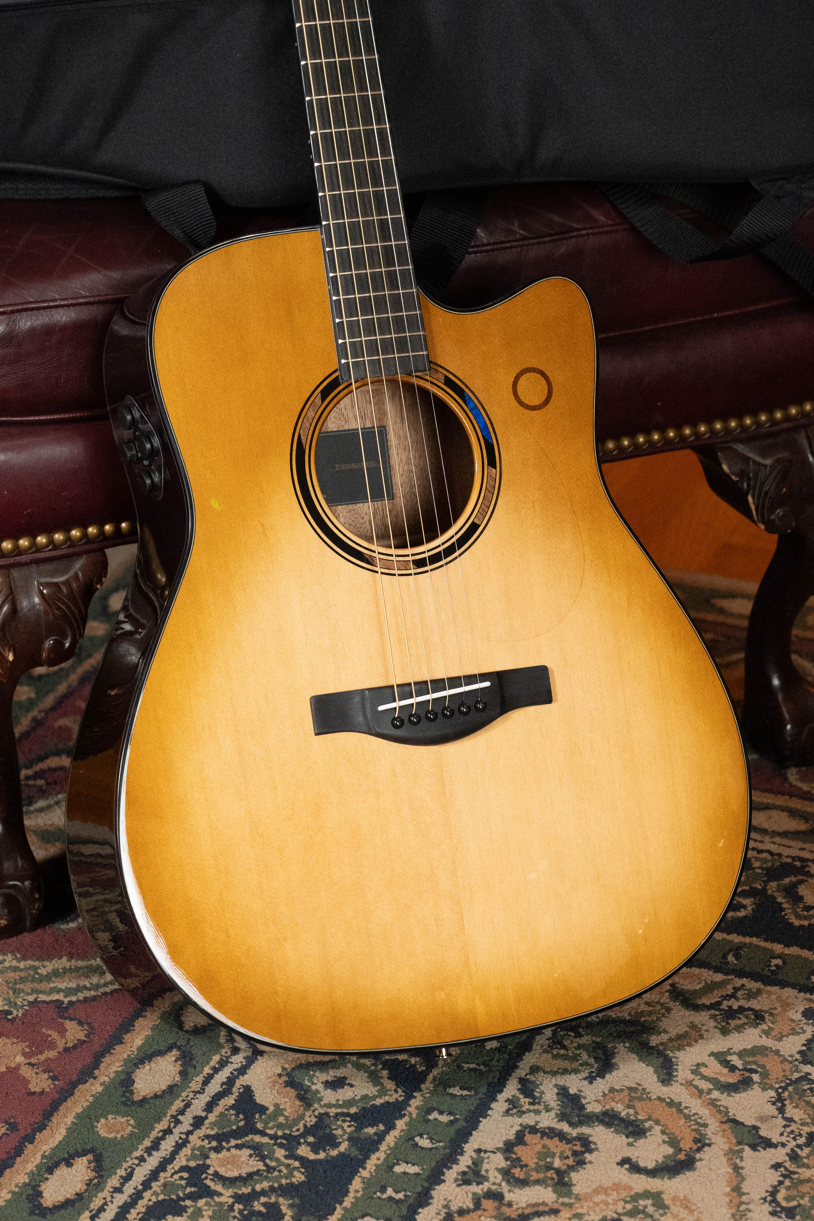 The Yamaha TAG3C-SDB Sitka/Mahogany Sandburst dreadnought guitar, featuring the NEWEST TransAcoustic System (#0688), rests against a dark red bench with brass studs on a patterned rug, highlighting its immersive sound capabilities.