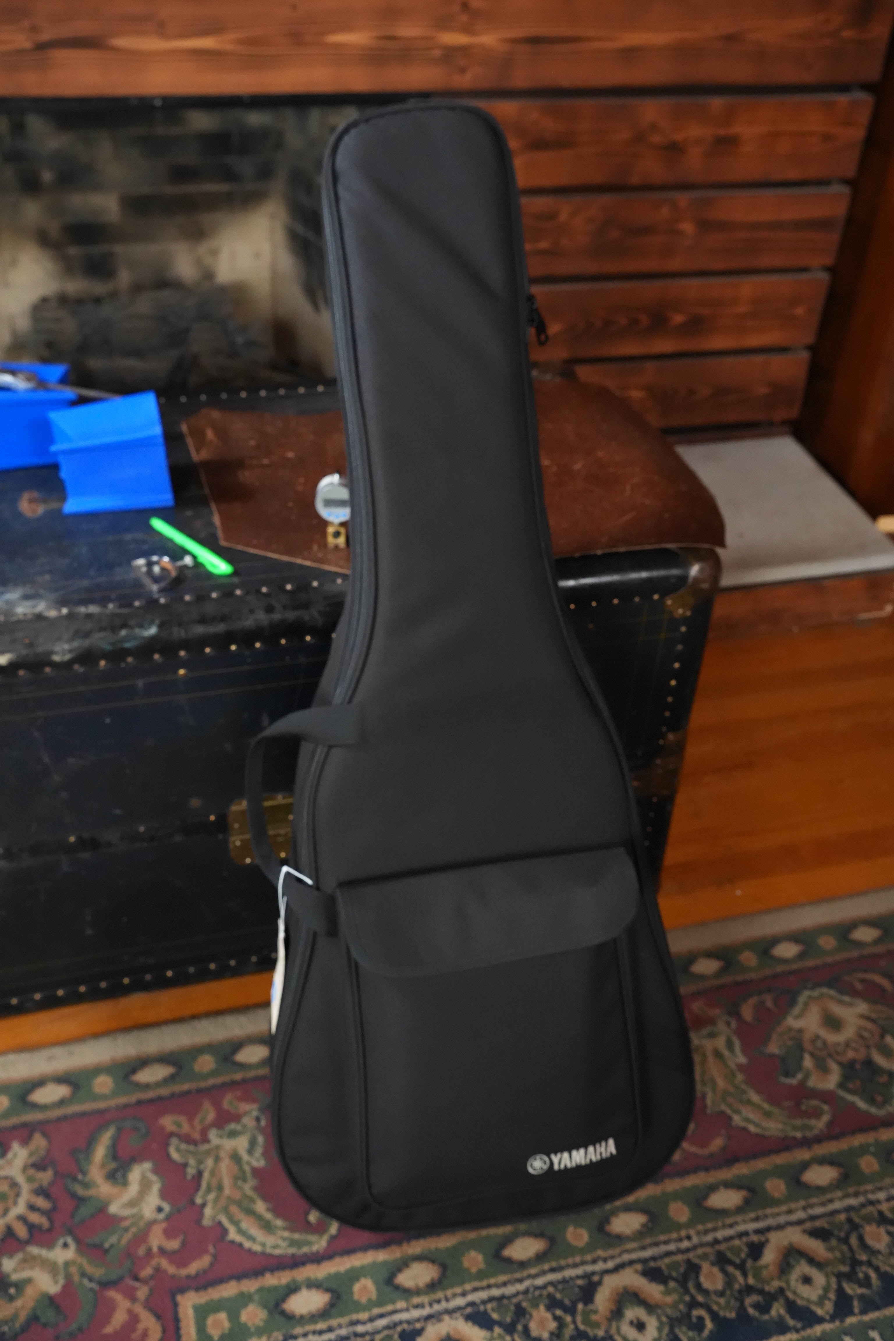 A black Yamaha gig bag stands upright on a patterned rug before a vintage trunk and wood wall, ideal for protecting your Yamaha TAS1 C Sitka/Mahogany Sand Burst Transacoustic Concert Guitar w/Cutaway #0132.