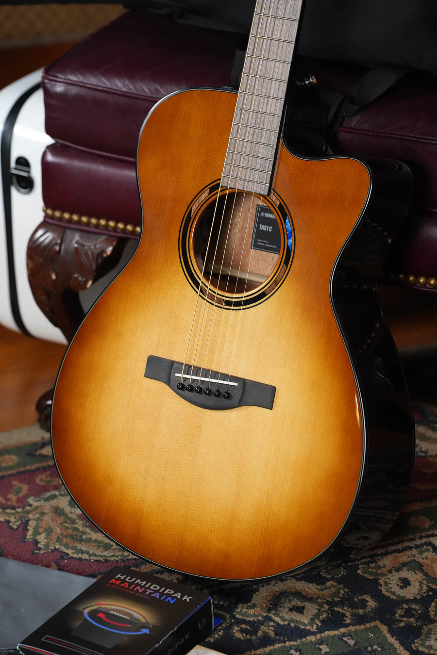 A Yamaha TAS1 C Sitka/Mahogany Sand Burst Transacoustic Concert Guitar w/Cutaway #0132 rests upright against a leather bench, beside the book "Mundira Muraki" on a patterned rug. A white guitar case is partly visible in the background.