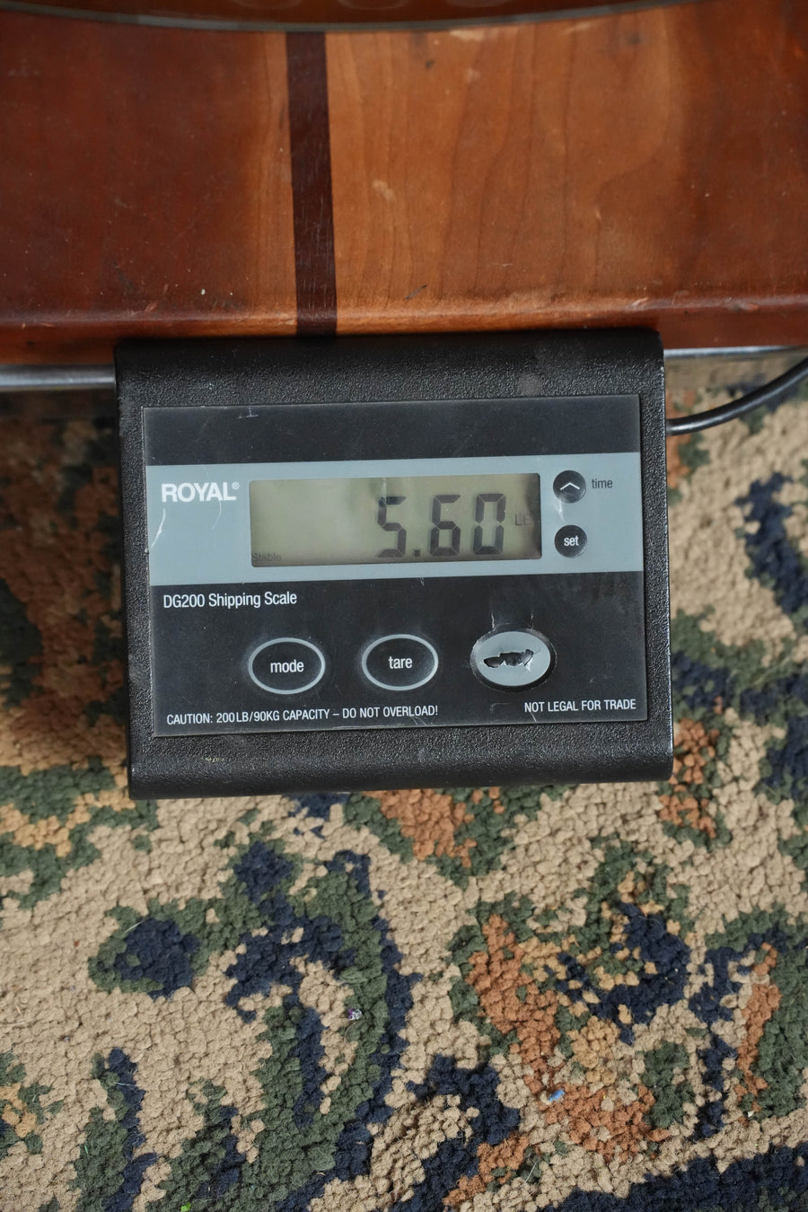 A Yamaha TAS1 C Sitka/Mahogany Sand Burst Transacoustic Concert Guitar w/Cutaway #0132 sits on a digital shipping scale reading 5.60 lbs. The scale, on a patterned carpet, shows mode, tare, on/off buttons, and a 200 lb max capacity.