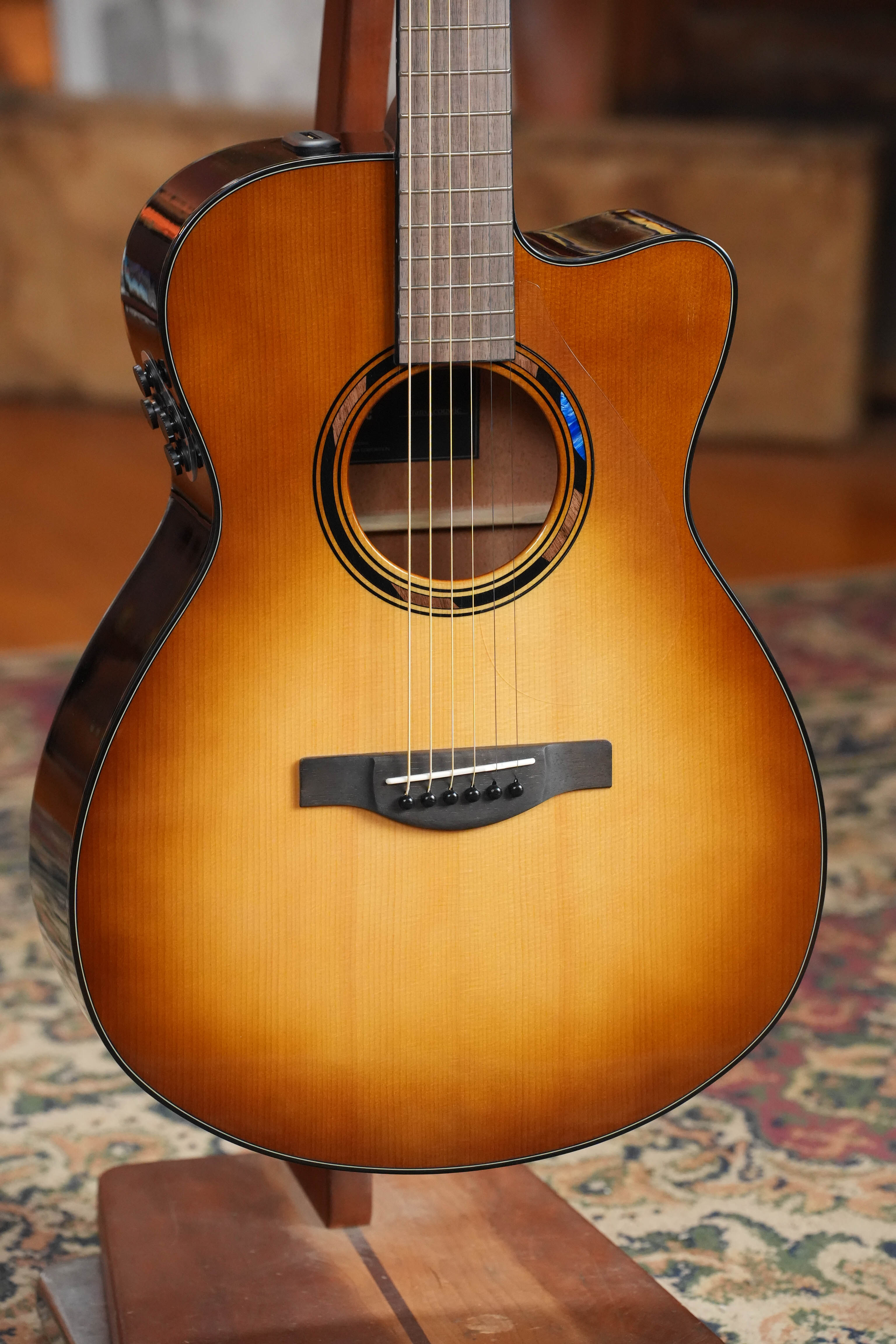 A close-up of a Yamaha TAS1 C Sitka/Mahogany Sand Burst Transacoustic Concert Guitar with cutaway (#0132) is displayed on a wooden stand over a patterned rug.