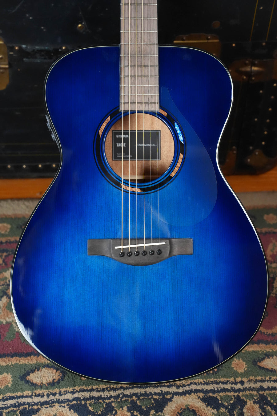 A Yamaha TAS1E Ultra Marine Spruce/Mahogany Transacoustic Concert Guitar #1161 with a glossy blue finish lies on a patterned rug, displaying its body, sound hole, bridge, fretboard, and advanced TransAcoustic system.