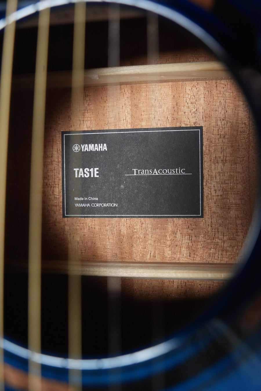 Close-up through the sound hole of a Yamaha TAS1E Ultra Marine Spruce/Mahogany Transacoustic Concert Guitar #1161, showing the model label TA51E and interior wood, with guitar strings visible in front.