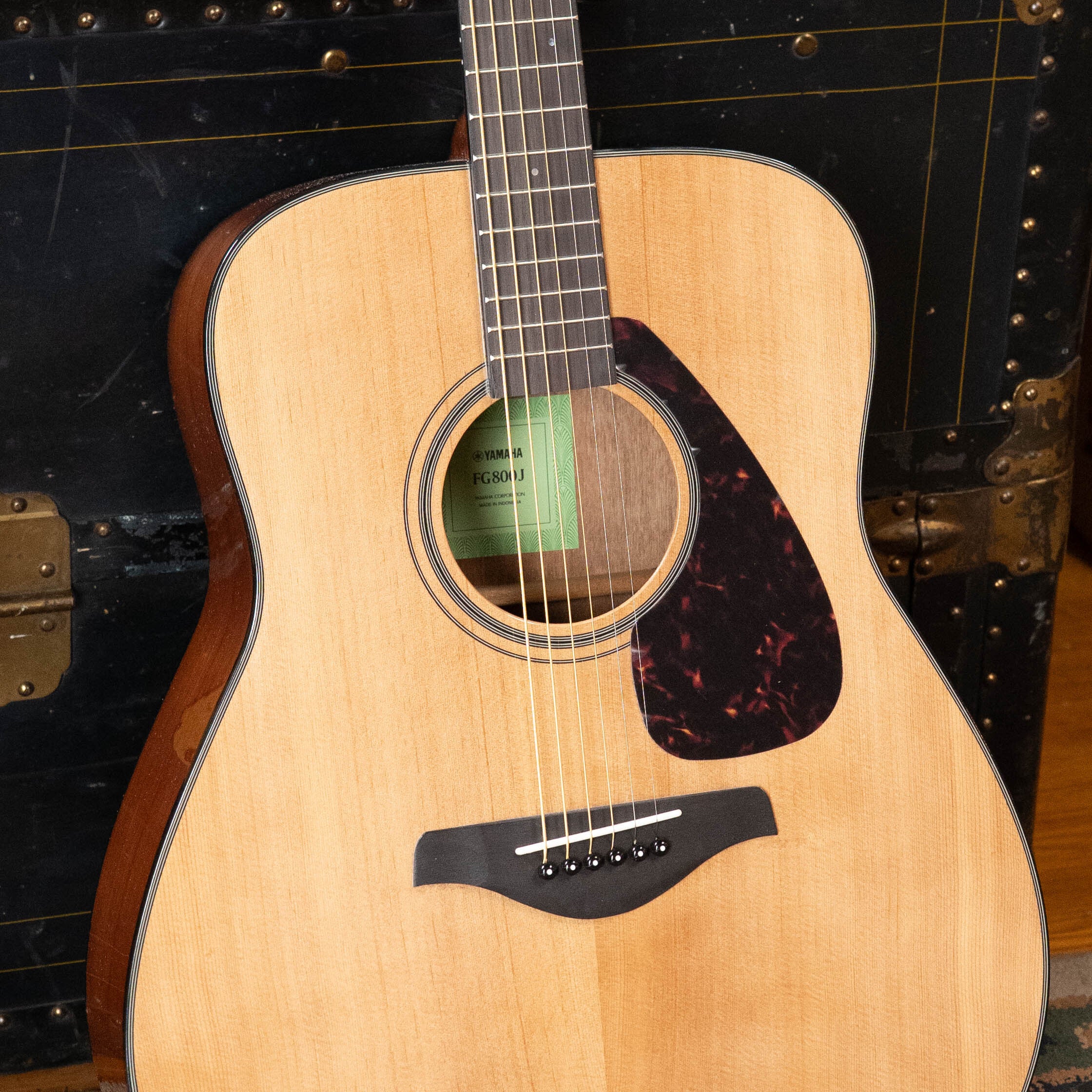 A close-up of the Yamaha GigMaker Deluxe Acoustic Pack in natural finish, featuring a light wood body and dark pickguard, resting before a black vintage trunk—an ideal starter acoustic guitar from Yamaha.