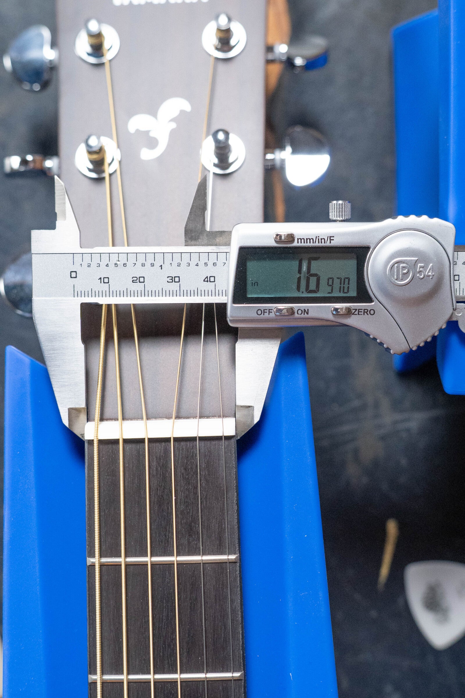 A Yamaha GigMaker Deluxe Acoustic Pack (Natural Finish #8229) guitar neck at the nut measures 1.6970 inches wide, as shown on a digital caliper. The dreadnought is held by blue supports, with its strings and headstock visible.