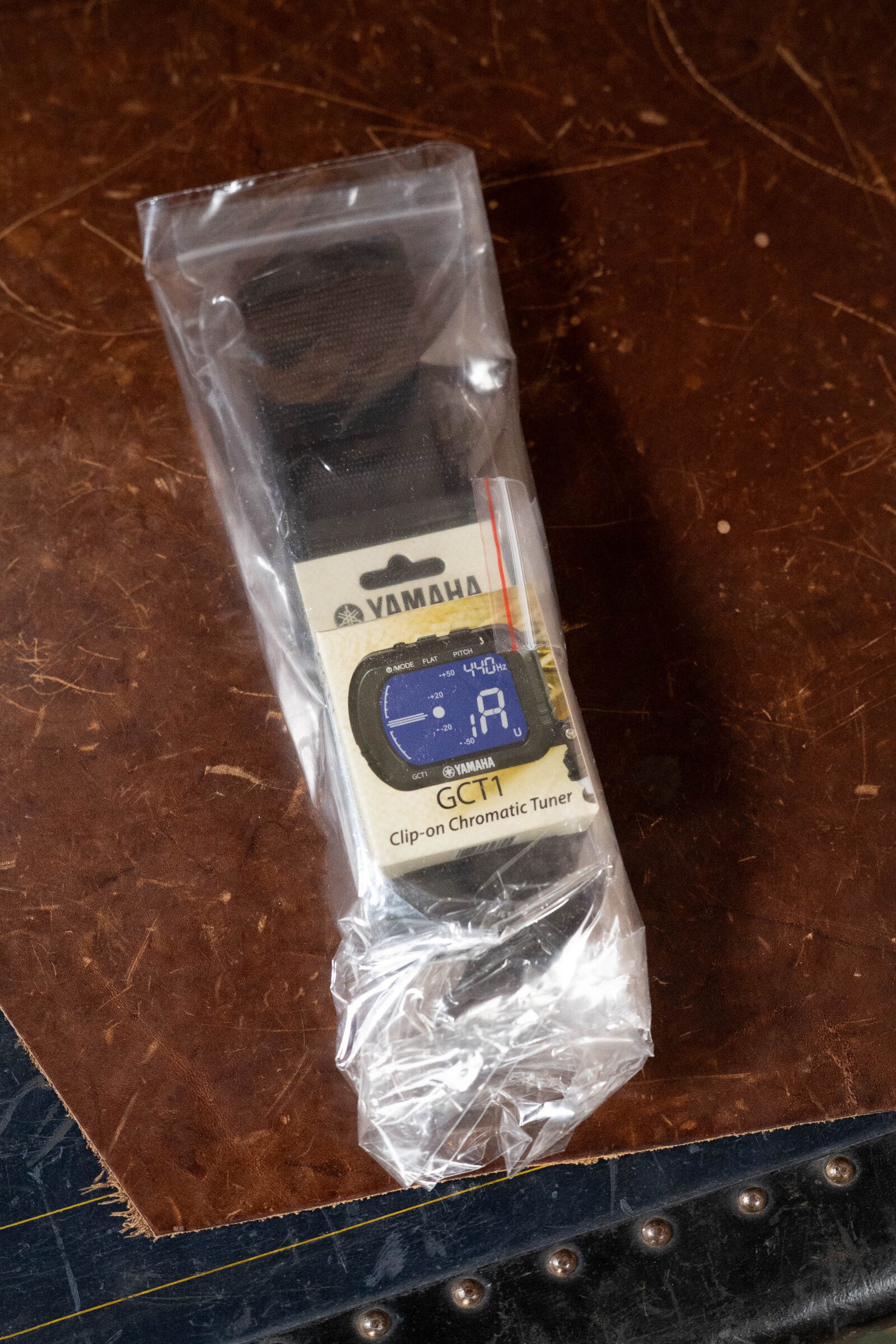 A Yamaha GCT1 clip-on chromatic tuner, still in its packaging and partially wrapped in clear plastic, rests on brown leather—an ideal accessory for the Yamaha GigMaker Deluxe Acoustic Pack - Natural Finish #8229.