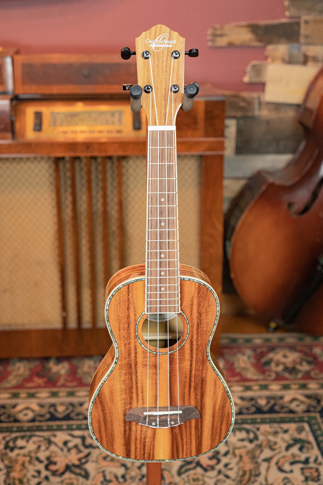 The Oscar Schmidt OU5K-A-U KOA Professional Concert Ukulele stands upright on a stand, showcased with vintage musical equipment and a patterned rug, featuring a glossy Hawaiian Koa finish.