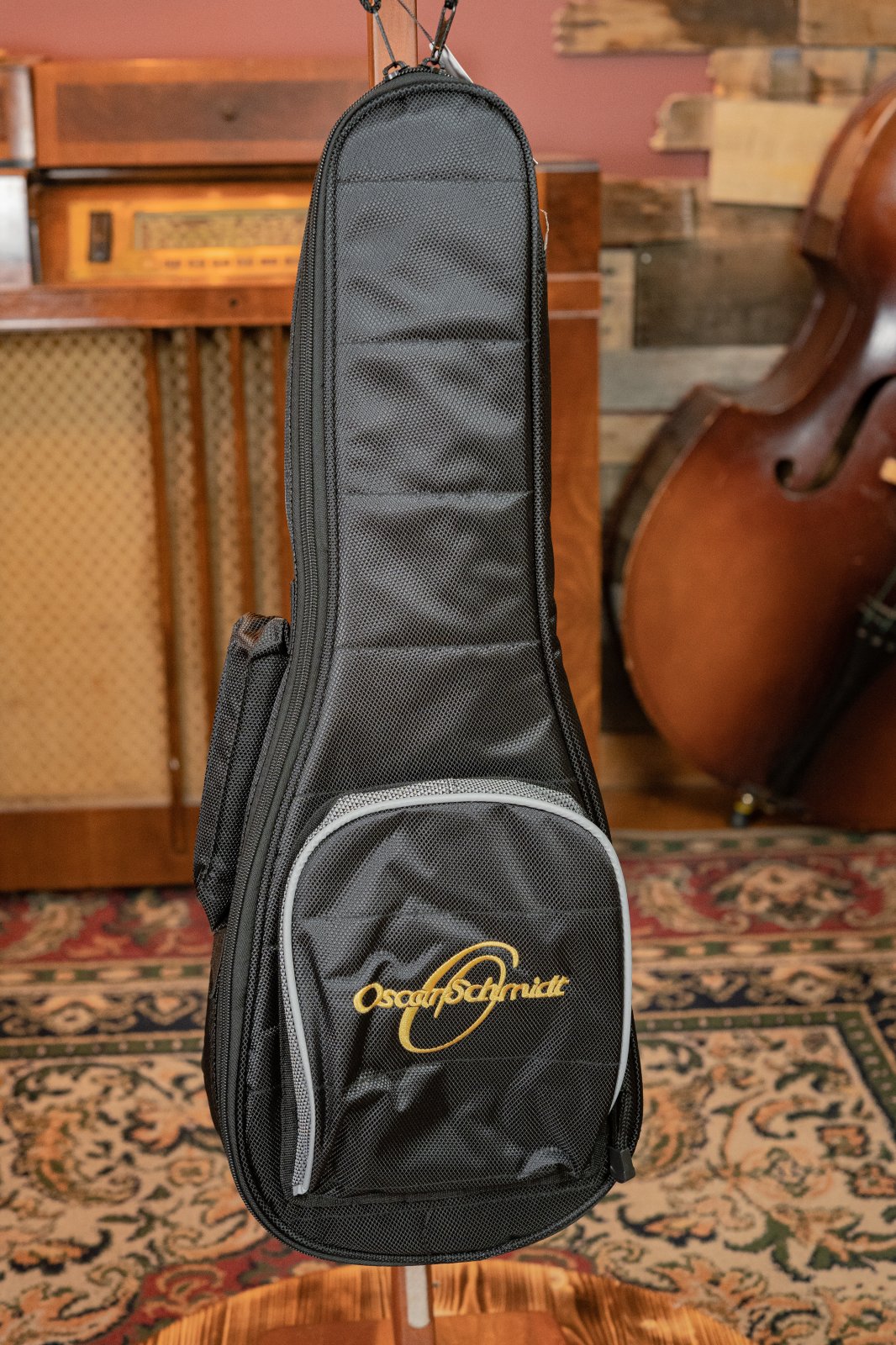 A black Oscar Schmidt gig bag with gold and white logo sits in a vintage-inspired room—complete with upright bass and patterned rug—the ideal home for your hand-crafted Oscar Schmidt OU5K-A-U KOA Professional Concert Ukulele.