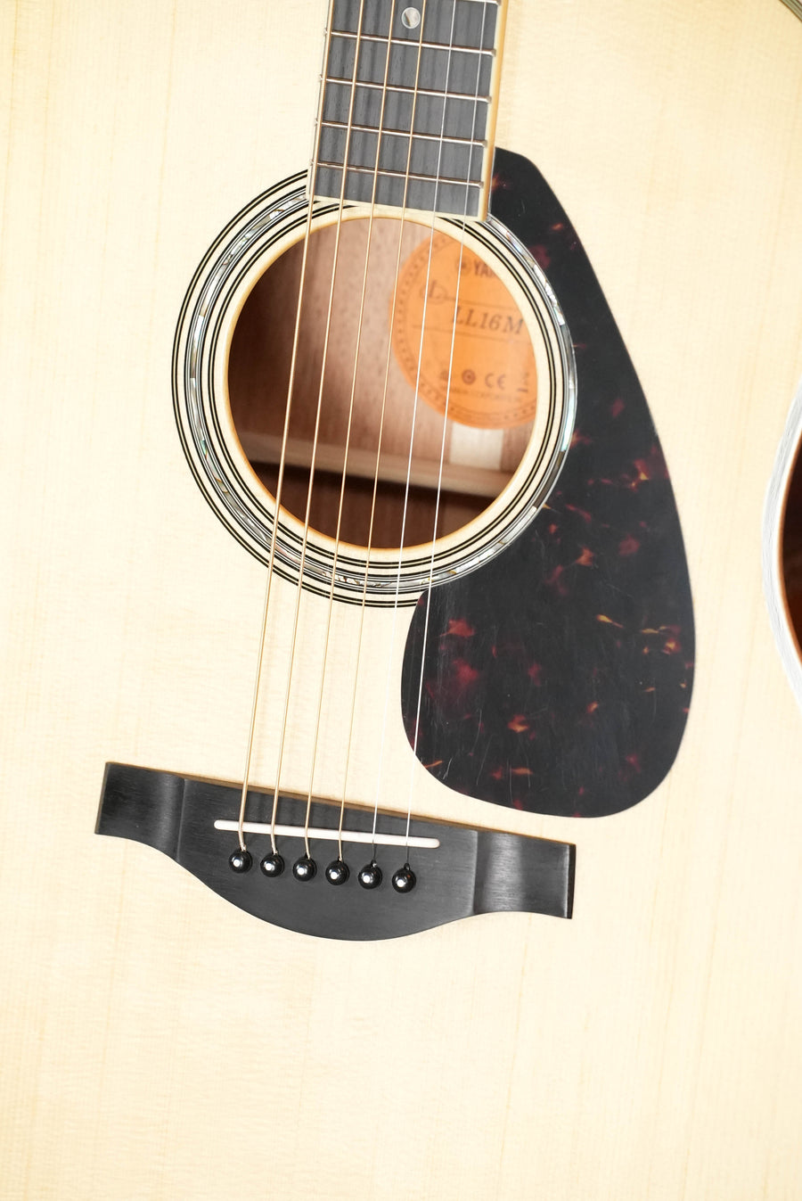 Close-up of a Yamaha LL16M ARE Engelmann/Mahogany Original Jumbo Acoustic Guitar with SRT Zero Impact Pickup, focusing on the sound hole, strings, bridge, and black pickguard on a light wood body.