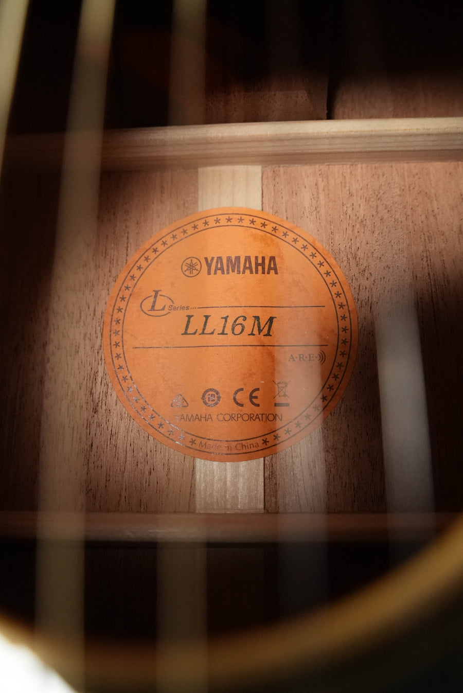 Close-up of a Yamaha LL16M ARE Engelmann/Mahogany Original Jumbo Acoustic Guitar label inside the soundhole, showing model details, A.R.E. process info, and Yamaha branding against a wooden background.