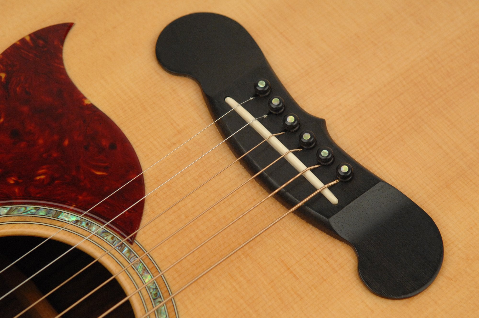 Close-up of six strings anchored by D'Addario Ebony Bridge Pins w/End Pin Set - Abalone Inlay and a wooden bridge, with an abalone rosette and patterned red pickguard on a light wood acoustic guitar body.